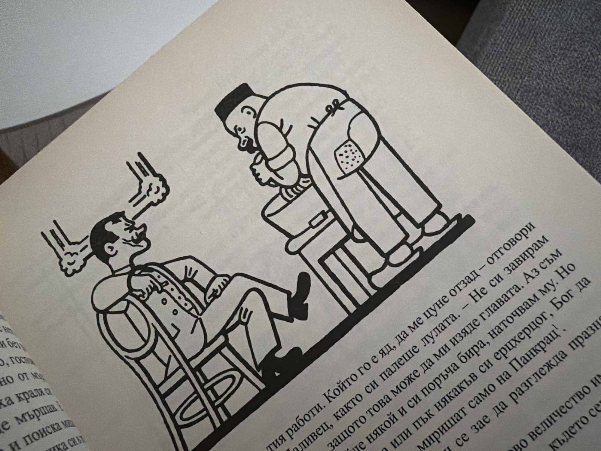 The image shows a black and white illustration of two cartoonish figures interacting. The figure on the left is larger and is seated in what appears to be a wheelchair or a similar contraption with large wheels. This figure is wearing a suit and hat and is leaning forward, with their arms extended. The figure on the right is smaller and is seated, holding onto an object that resembles a bucket or a large container. This figure is wearing a hat and a button-up shirt, with a detail on their shorts that looks like a polka dot pattern. The illustration depicts an action, possibly a conversation or a dispute, with animated expressions on both figures' faces. There is text in Bulgarian on the page below the illustration. The image is of a book open to a page with this illustration and text. There is no discernible context or location information related to the city of Pazardzhik, Bulgaria within the image itself.