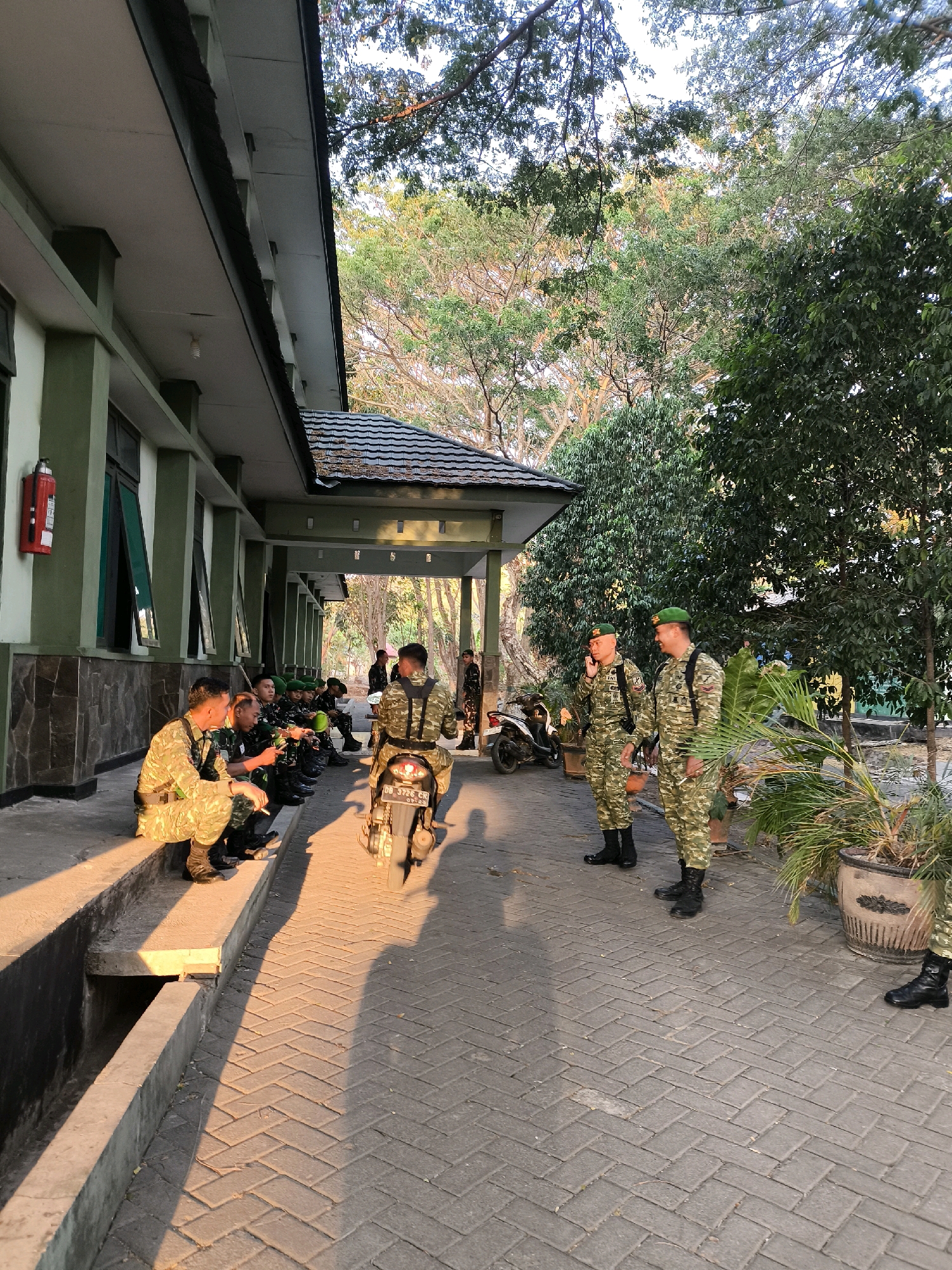 In an outdoor setting, several individuals in military camouflage uniforms are present. The scene appears to be in front of a building with a green facade, a covered walkway, and trees in the background. The lighting suggests it is either early morning or late afternoon, with long shadows cast on the brick-paved ground.
In the foreground, a group of soldiers are seated on a concrete ledge along the building. One soldier is actively smoking a cigarette. To the right, another soldier is seated on a motorcycle. Further to the right, two more soldiers stand, one of whom is engaged in a phone conversation. The motorcycle has a license plate that reads "DB 3726 CB". The building has several windows, and a red fire extinguisher is mounted on the wall. Potted plants are visible to the right, near the standing soldiers. The overall environment suggests a military compound or barracks.