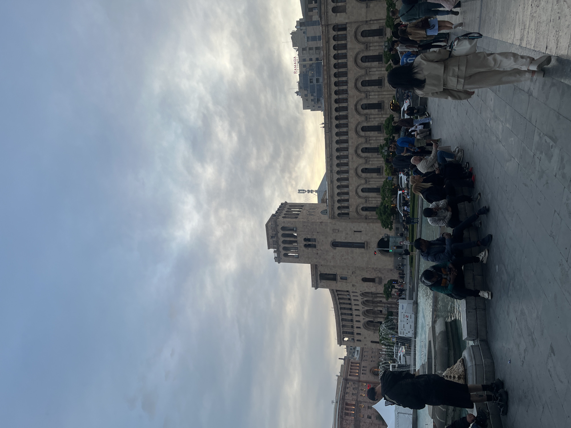 The image captures a scene in Yerevan, Armenia, featuring the Republic Square. Several buildings with arched windows and classical architectural details form the backdrop. A large group of people is present, some seated along the edge of a fountain area, others standing and walking. The sky is overcast with clouds, and the light is diffused, suggesting either early morning or late afternoon. Some people appear to be tourists. The ground is paved with stone. A building with the word "RAMADA" can be seen in the background.