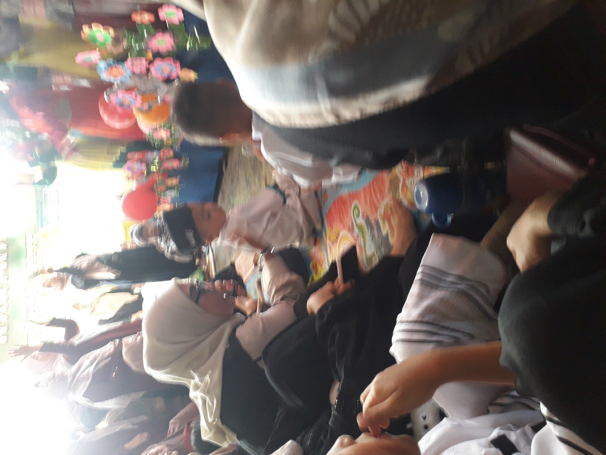 The image depicts a dimly lit indoor gathering, possibly a celebration or event, with several individuals present. In the foreground, a person wearing a white hijab and glasses is visible, looking to the right. To their left, another individual is partially visible, also wearing a hijab. A young child in a white outfit and a dark cap is seated in the middle ground, facing away from the camera. Behind the child, another individual in a white shirt is also visible. The background features a table adorned with colorful decorations, including balloons and what appear to be paper flowers. The floor is covered with a patterned mat. The overall atmosphere suggests a community event, possibly in Cangkung, Indonesia, given the context provided.