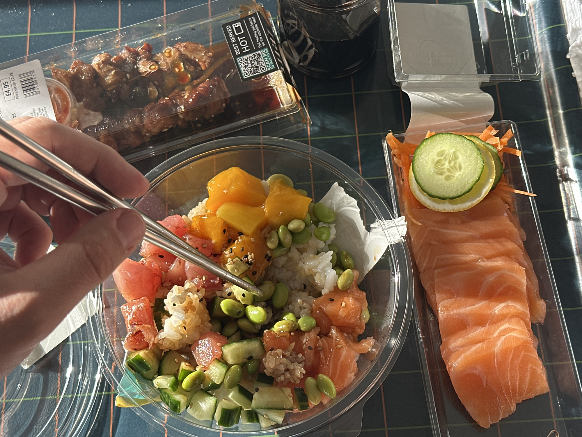 The image depicts a person using chopsticks to eat a meal. The meal consists of a poke bowl with rice, salmon, tuna, mango, cucumber, and edamame. There is also a container of salmon sashimi with cucumber and lemon slices, a container of meat skewers, and a beverage. The packaging indicates that the food was purchased in London, United Kingdom. The environment appears to be a dining area.