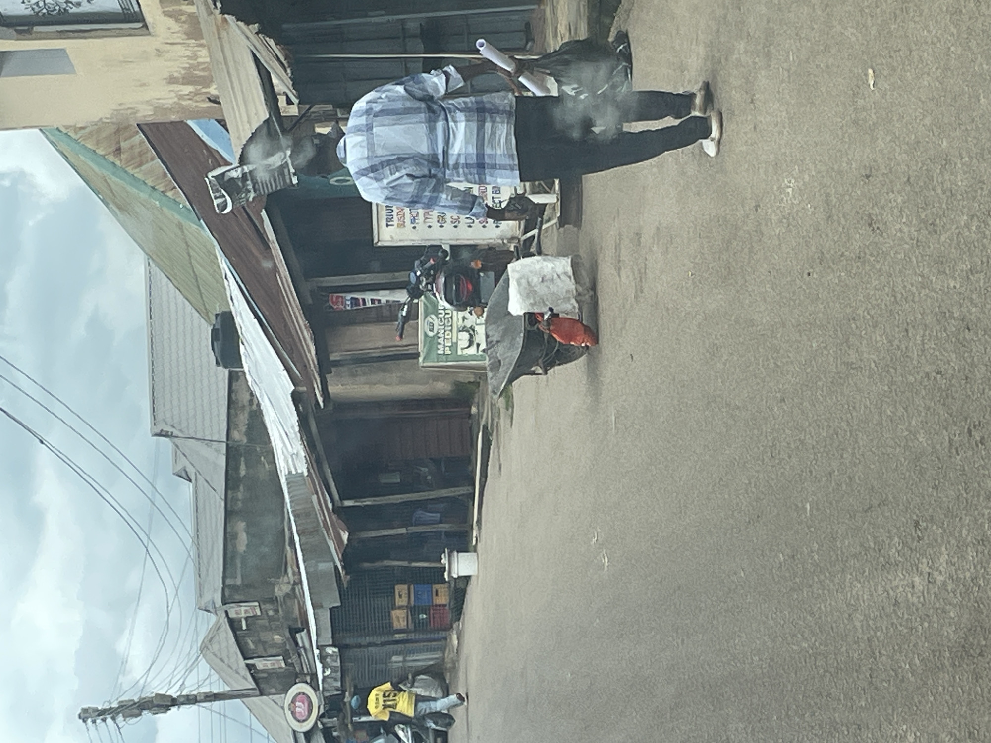 The image depicts a street scene in Jos, Nigeria, characterized by weathered buildings and a cloudy sky. A man in a blue and white checkered shirt and black trousers is walking away from the camera, carrying a roll of paper and a black plastic bag. Another person, wearing a yellow shirt with "115" on the back and a blue cap, is walking away from the camera on the left side of the street, carrying a black plastic bag. A motorcycle is parked behind a wheelbarrow on the right side of the street. Various market stalls and shops with corrugated iron roofs line the street, with stacked crates visible in front of one of them. Electrical wires are strung across the sky, supported by a utility pole. The overall impression is one of a bustling, somewhat impoverished urban environment.