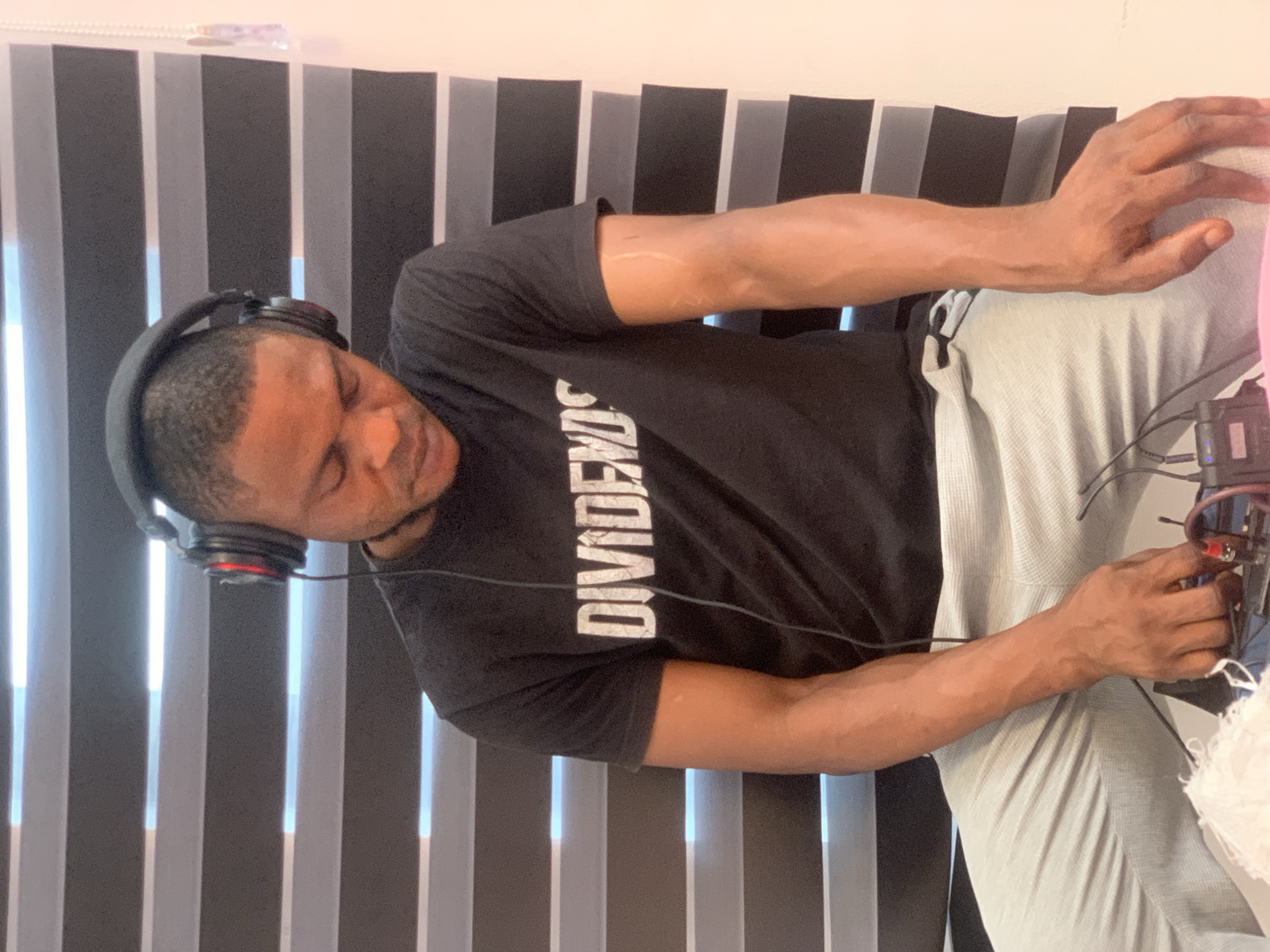 An indoor scene in Lagos, Nigeria, features a dark-skinned adult male seated. He is wearing a black t-shirt with "DIVIDENDS" printed in white across the chest, and light grey textured pants. Over-ear headphones, dark with red accents on the earcups, are worn on his head, with a cable extending downwards in front of his torso. His head is tilted downwards, and his gaze is directed intently at a small black electronic device in his lap, which is surrounded by multiple wires and a crumpled piece of white fabric. His hands are positioned over this device, appearing to manipulate it. Prominent veins are visible on his forearms, and a distinct linear light-colored scar is present on his right forearm. The background consists of horizontal blinds with alternating dark and illuminated light strips, suggesting a window behind them. A plain, light-colored wall is visible to the right of the blinds, which also display a translucent pulling mechanism on their far right edge.