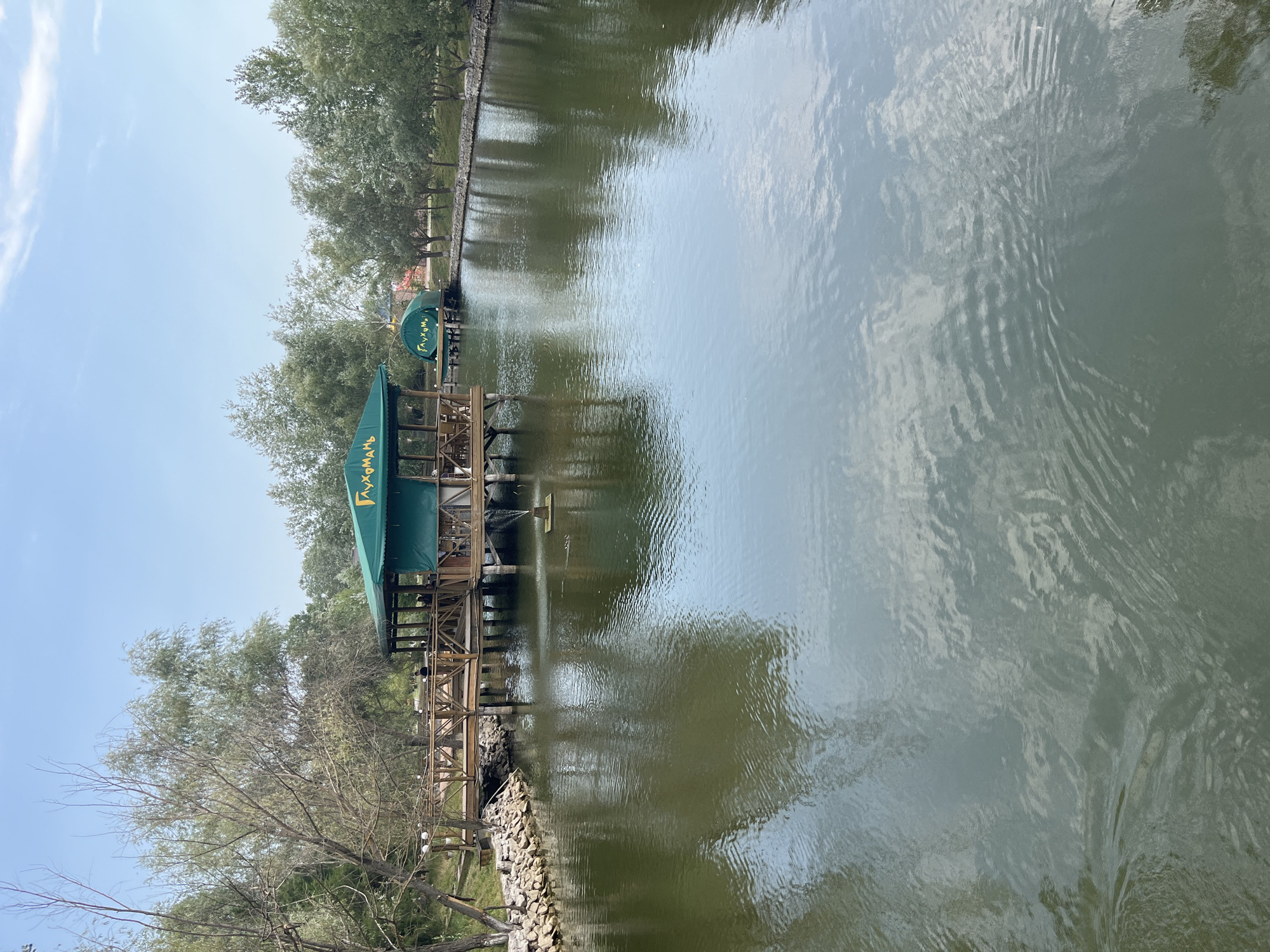 The image shows a picturesque view of a calm body of water in Shcherbani, Ukraine, with a large wooden pier extending into the water. On the pier, there is a green-roofed structure, possibly a restaurant or cafe, with text in Cyrillic script that reads "Глухом", which translates to "Deaf". The structure has tables and chairs visible, and a few people are seated there. Further in the background, a smaller green structure is also visible on the water, similar in design to the larger one. A small fountain is active in the water near the pier, creating ripples. The water is surrounded by lush green trees and vegetation, and the sky above is clear with a few wispy clouds. The overall scene conveys a serene and tranquil atmosphere.