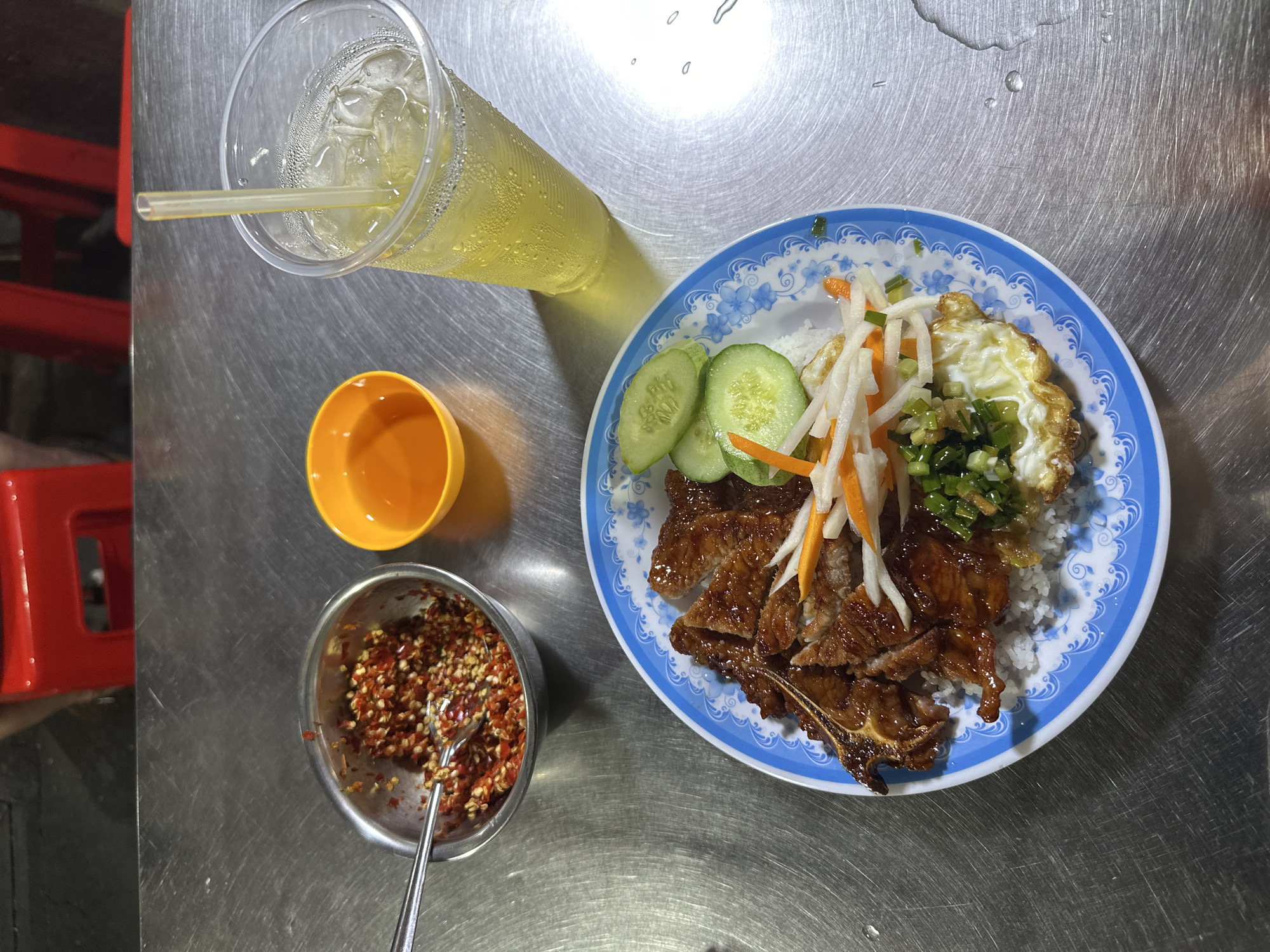 An overhead shot captures a casual meal set on a brushed metal table, likely at a Vietnamese street food stall or local eatery. The main focus is a plate of food, a drink, and condiments.
The central dish features a generous portion of white rice topped with slices of glossy, grilled marinated meat (resembling pork chops or ribs, typical of *cơm tấm*), a fried egg with a slightly crispy edge, and a garnish of pickled daikon and carrot (*đồ chua*). Sliced fresh cucumber and chopped green onions drizzled with oil (mỡ hành) are also visible on the plate, adding color and freshness. The plate itself is white with a distinctive blue floral and scroll pattern along its rim.
To the left of the plate, a tall plastic cup holds a light yellow iced drink with a straw, showing condensation on its exterior, indicating its coldness. Below the cup, a small, empty orange plastic bowl sits on the table. Further down, a small metal bowl contains a vibrant red mixture of chili flakes, possibly mixed with oil or a sauce, accompanied by a small metal spoon.
The metal table surface shows visible scratches and reflections from what appears to be artificial light, suggesting the meal is being consumed in the evening or at night. Several water droplets are scattered across the table surface, likely from the condensation of the cold drink. In the left background, part of a red plastic chair is visible, reinforcing the casual, local dining environment typical of Thủ Đức, Vietnam, where such meals are popular. There is no visible text in the image.