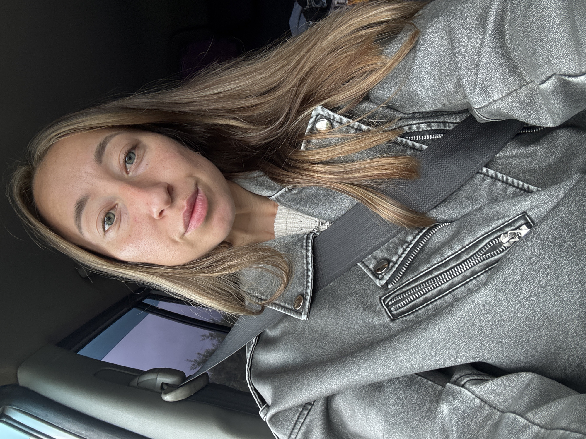 The image displays a head-and-shoulders portrait of a female individual seated within the passenger compartment of a vehicle, located in Novyi, Russia. The subject, positioned centrally, looks directly at the camera with a neutral expression. She has long, light brown hair with blonde highlights, a fair complexion with visible freckles across her nose and cheeks, and green eyes. Her lips are full and slightly parted. She is wearing a metallic grey faux leather motorcycle jacket, featuring multiple silver zippers and snap buttons on the collar and lapels, over a cream-colored knit sweater, visible at the neckline. A dark grey seatbelt is fastened across her chest, traversing from her left shoulder downwards. To her left, through the vehicle's window, distant trees and foliage are visible under a muted, possibly overcast or twilight sky, rendered with a purplish tint by the window glass. The interior of the car, including a light grey pillar on the left and dark elements in the background, is also discernible.