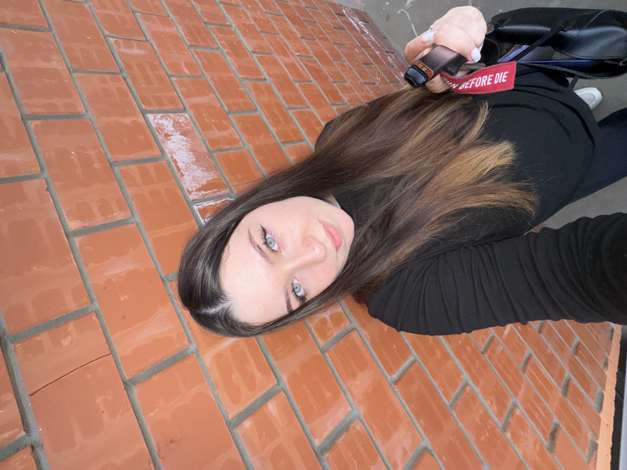 In a medium shot, a young woman with long, dark brown hair and light blue eyes stands against a brick wall. She is wearing a black long-sleeved shirt and black pants. She is holding a vape device with a red keychain attached, and a black bag is slung over her shoulder. The brick wall is orange-red and appears to be outdoors. The scene suggests an urban environment, potentially Perm, Russia, based on the commonality of brick architecture in the region.