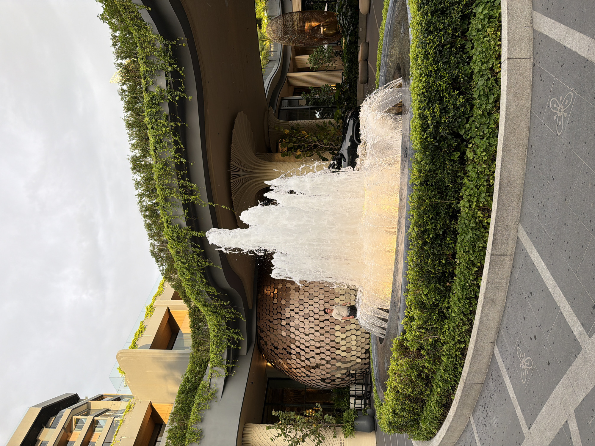 A fountain is the focal point of the image, with a person standing near its base. The surrounding environment includes a building with a unique architectural design, featuring greenery covering its exterior. The ground is paved with gray tiles marked with a white butterfly motif. The location is identified as Pattaya City, Thailand.