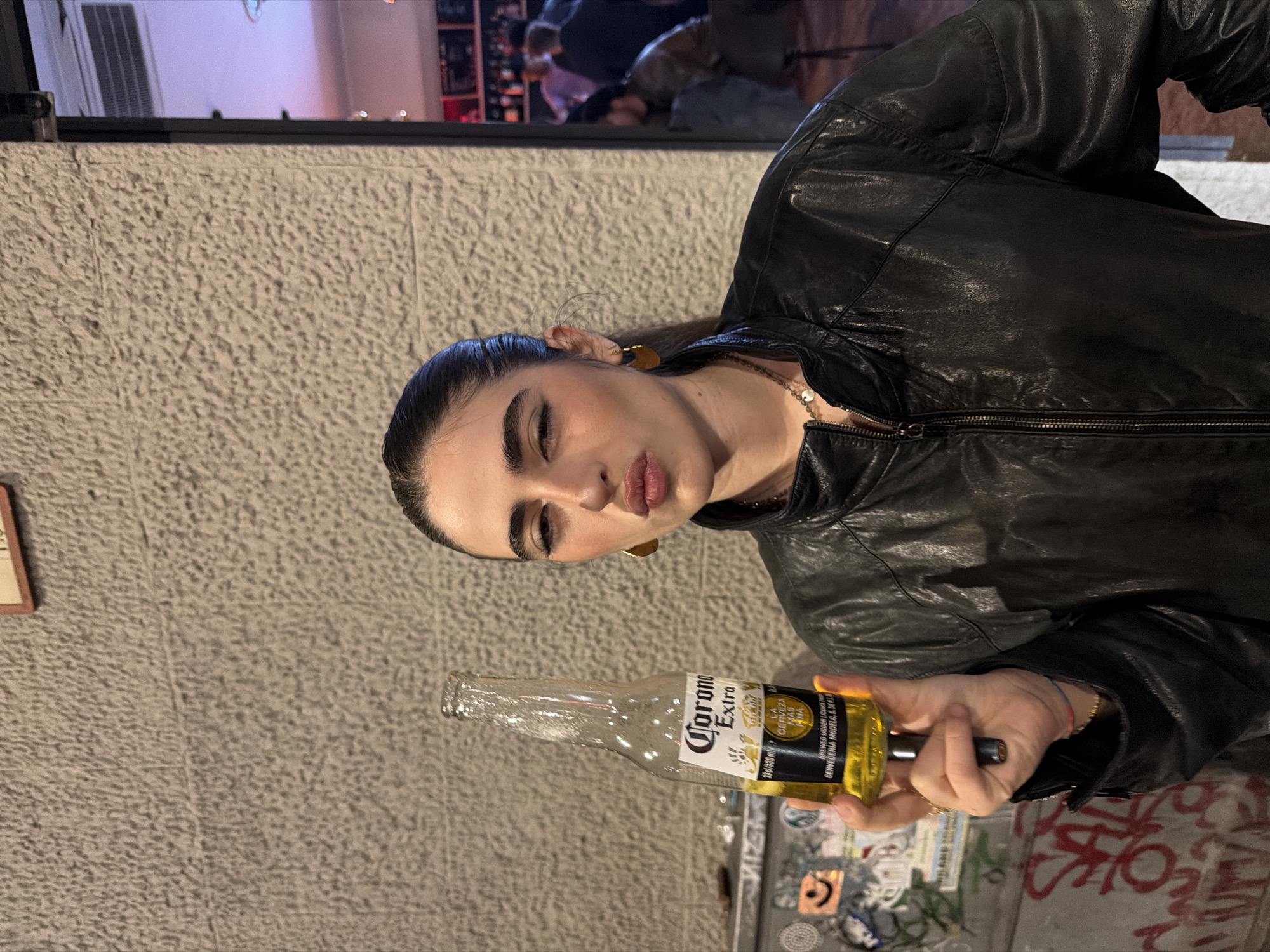 A woman in a black leather jacket is holding a bottle of Corona Extra beer. The background shows a textured wall and part of a bar or restaurant scene with other people present. The location is possibly in Milan, Italy. The environment appears to be an urban setting, likely a nightlife area in the city. The image depicts a casual scene, possibly at a bar or outdoor setting.