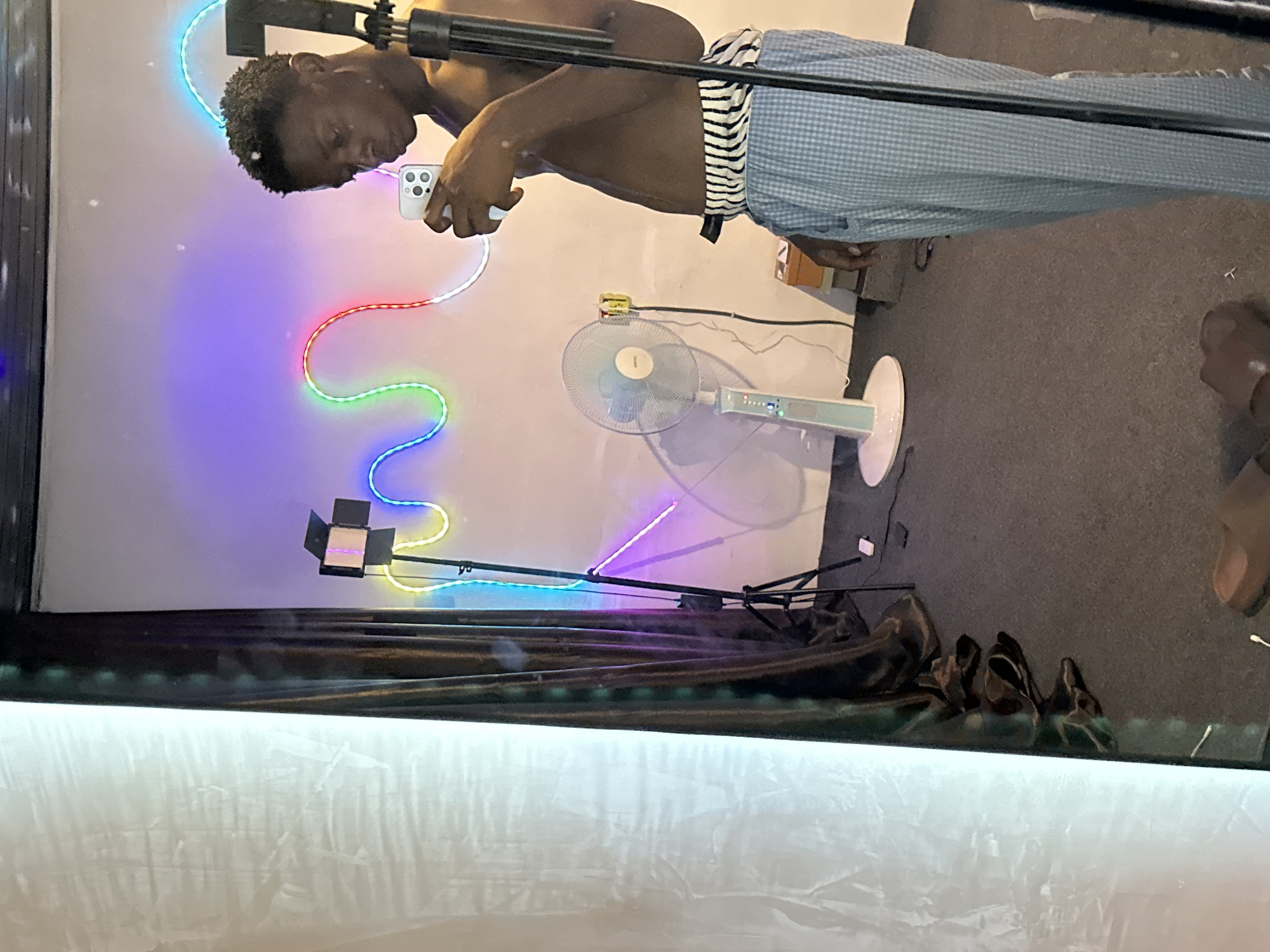 A man stands indoors, shirtless and wearing patterned pants. He holds a smartphone in his hand and appears to be filming or taking a picture of a colorful LED light installation. A small studio light on a stand, a rotating fan, and decorative objects are visible in the background. The room's floor is carpeted. The location is not specified within the image and cannot be determined from the image. No information suggests this is in Bukuru, Nigeria.