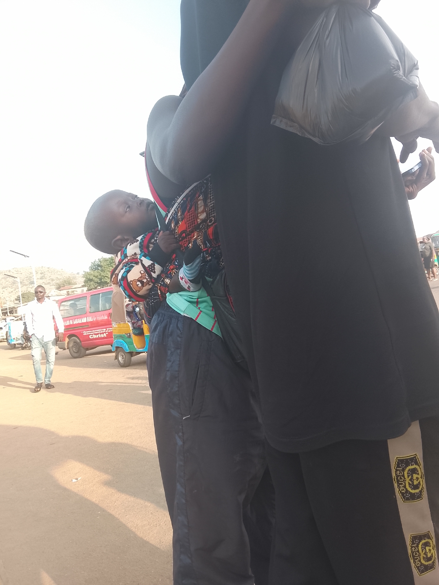A medium shot reveals a person in Gwong, Nigeria, carrying a young child, with a man walking in the background.
The person carrying the child is wearing dark clothing. The child is wrapped in a colorful patterned carrier. A black plastic bag is visible on the person's shoulder.
In the background, a man in a white shirt and jeans walks on a street with a red van and a tricycle. The setting appears to be a street scene.
