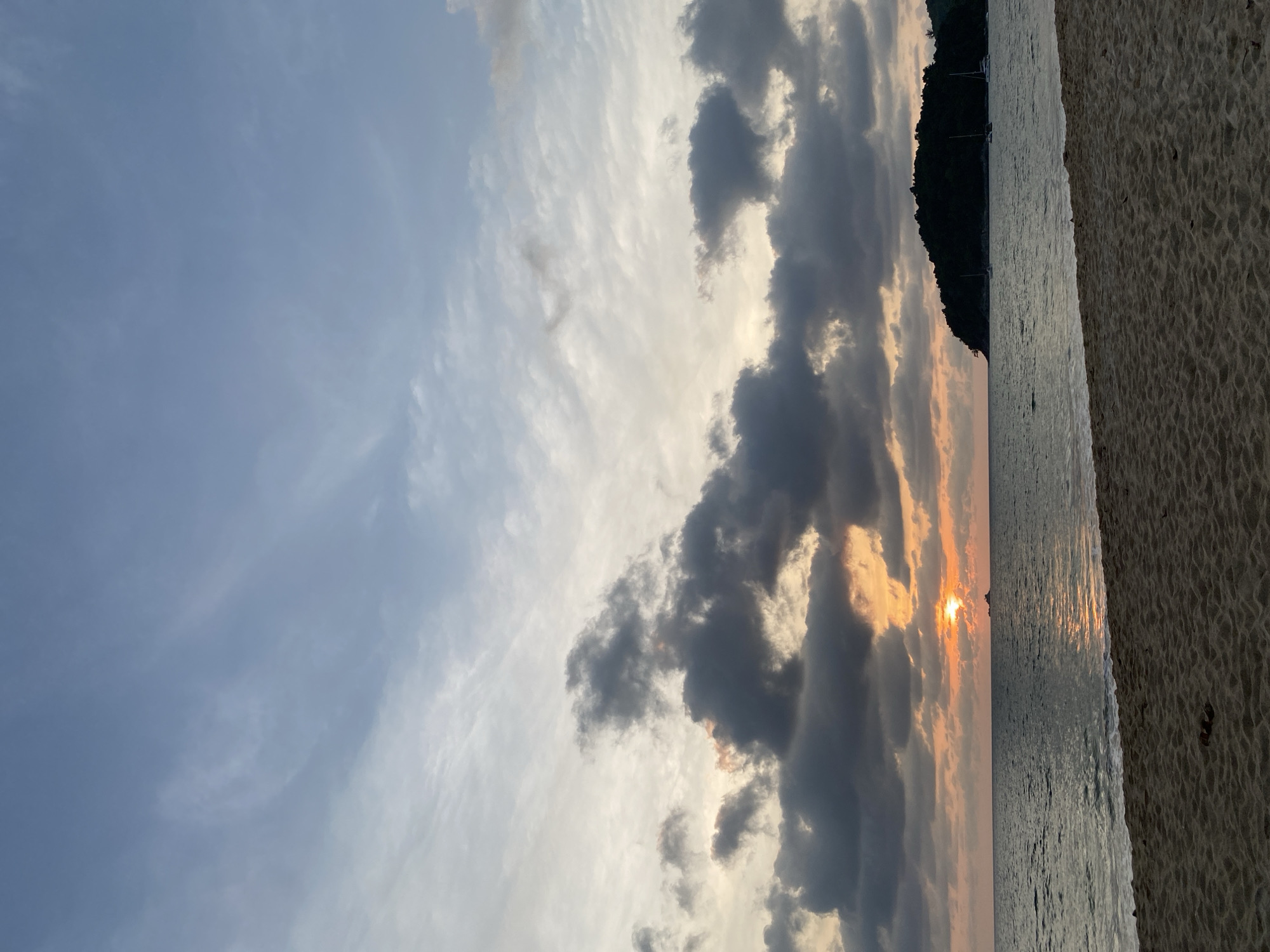 The image captures a serene coastal landscape at either sunrise or sunset, set against the backdrop of Choeng Thale, Thailand. The view is composed of sky, sea, a distant landmass, and a sandy beach in the foreground.
The upper portion of the frame is dominated by a dynamic sky. Patches of clear, light blue are interspersed with various cloud formations. There are delicate, wispy white clouds stretching across the sky, as well as heavier, darker grey clouds. These darker clouds are dramatically lit from behind by the low-lying sun, creating warm hues of orange, yellow, and subtle pinks around their edges and undersides.
The sun, a brilliant orb, is visible near the horizon, partially obscured by the clouds. Its intense glow casts a shimmering path of light across the calm water surface, which reflects the varying light conditions of the sky. In the mid-ground, across the water, a dark, silhouetted landmass, likely a hilly island or a mountainous coastline densely covered in foliage, stretches along the horizon. Several faint, indistinct shapes, possibly small boats, can be seen on the water near this landmass.
The immediate foreground features a sandy beach, textured with subtle ripple patterns left by the receding tide or waves. The overall atmosphere is peaceful and scenic, emphasizing the natural beauty of the coastal environment during a transition period of the day. No discernible text is visible in the image.