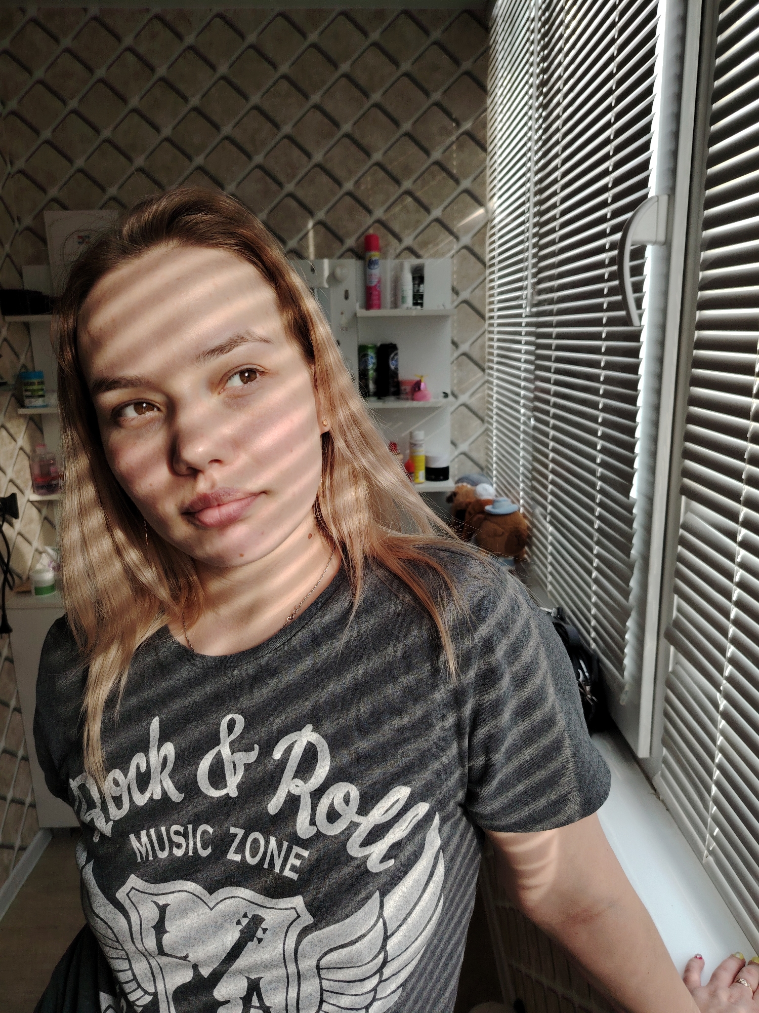 A female individual is depicted in a medium close-up, taken indoors within what appears to be a residential room in the city of Марковский, Russia. She has light brown hair, brown eyes, and a light complexion, with striped shadows from window blinds cast across her face and torso. She wears a dark grey short-sleeved t-shirt with a white graphic text reading "Rock & Roll MUSIC ZONE" above a design of a guitar and wings. A thin gold-colored necklace is visible. Her left hand, featuring multi-colored nail polish and a ring on the ring finger, rests on a white windowsill to the right. To her right, a white window frame with horizontal blinds, through which sunlight is entering, is prominently visible. The background to her left features a wall with textured, patterned wallpaper and white wall-mounted shelves holding various containers, including a pink aerosol can. A brown stuffed animal is visible on a surface near the window or shelves.