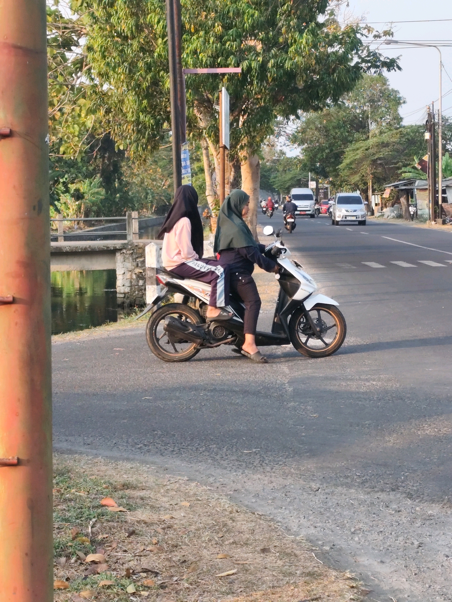 Two individuals, identified as females by their attire, are seated on a white scooter on the side of a paved road. The person in front, wearing a black hijab and a pale pink top with striped pants, is looking towards the right. The person behind, wearing a green hijab and a dark blue outfit, is operating the scooter. They are positioned on the right lane of a two-lane road. In the background, there are trees and vegetation, a bridge over a body of water, and other vehicles including a white van and a silver car on the road. The scene appears to be in a rural or semi-urban area of Bambanglipuro, Indonesia, characterized by the presence of local flora and a modest road infrastructure. The lighting suggests it is daytime, likely late afternoon due to the warm tones in the image.