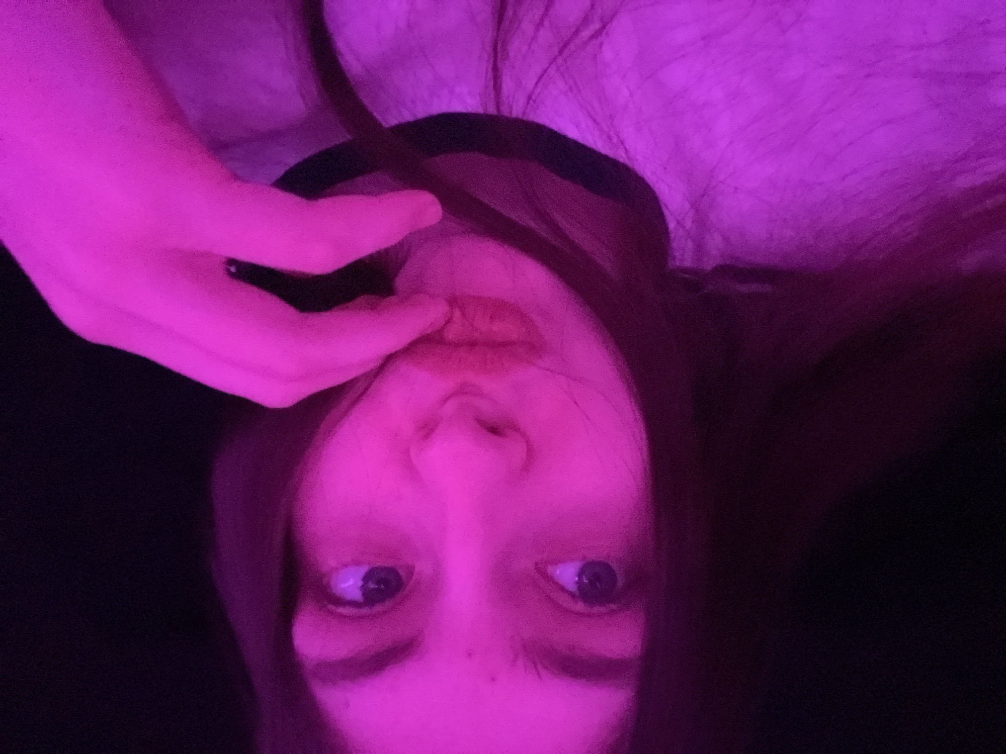 A close-up, upside-down selfie of a person illuminated by pink lighting. The person's face is central, featuring eyes looking up, a nose, and lips touched by fingers. The person has dark hair. The background is mostly dark except for a fuzzy pink area. The location is indeterminable.