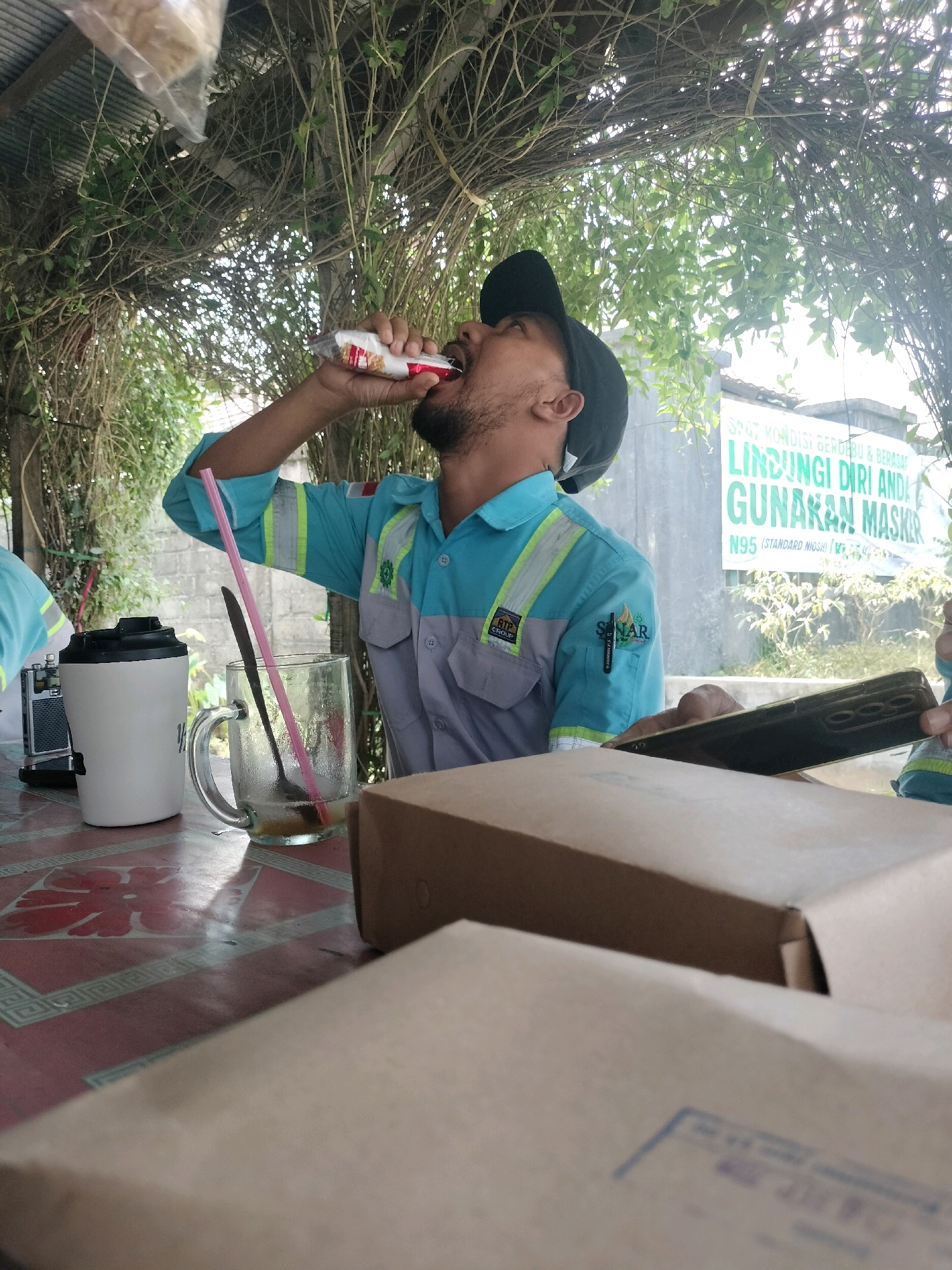 A man wearing a blue and gray work shirt and a black baseball cap is drinking from a packaged drink. He is seated at a table outdoors, with a clear glass of amber liquid and a white thermal mug on the table in front of him. Two brown cardboard boxes are also on the table, one in the foreground and one in the midground. In the background, a sign on a wall reads "SPOT, KONDISI BERDEBU & BERASAP - LINDUNGI DIRI ANDA - GUNAKAN MASKER - N95 (STANDARD NIOSH)". This suggests the location is Maburai, Indonesia, likely a work site or outdoor eating area. The man is holding the drink with his right hand, and his left arm is bent as he drinks. The scene is illuminated by natural light, with the sun filtering through the leaves of an overhead canopy of vines.