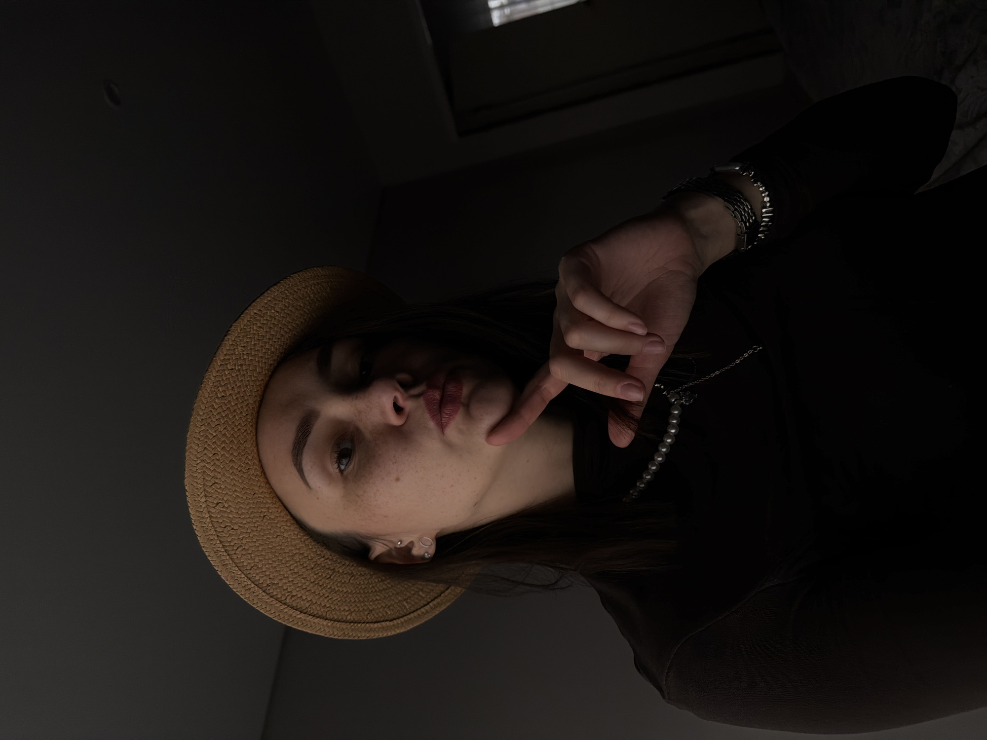 A woman is centrally positioned in the frame, wearing a light brown straw hat, a dark top, and jewelry consisting of bracelets and a pearl necklace. Her face is visible, showing freckles. Her hand is raised to her face. The background consists of a dark gray wall and ceiling. The scene is dimly lit. There is no indication of the image location being Chaikovsky, Russia.
