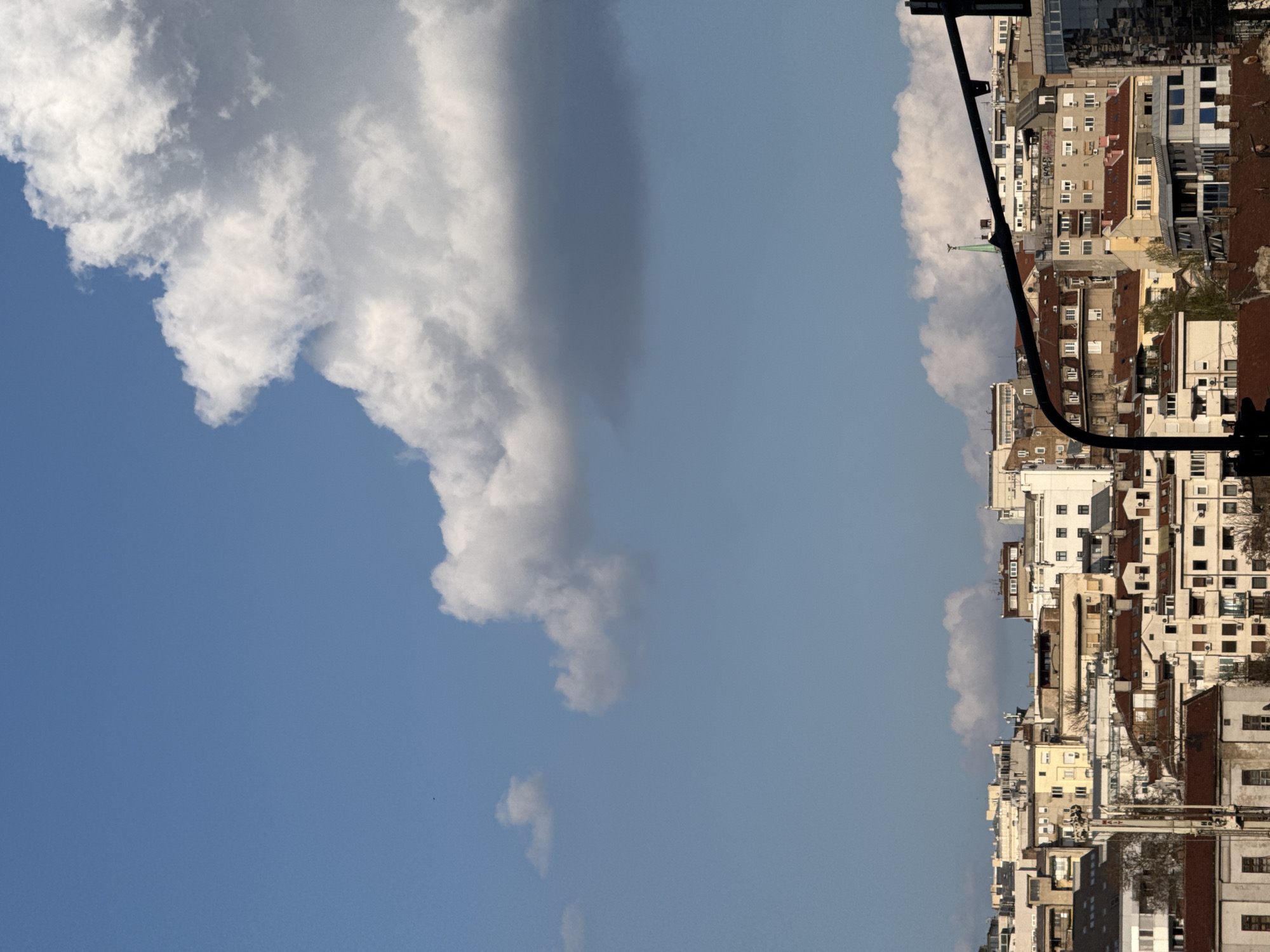 The image shows a view in Belgrade, Serbia, with a juxtaposition of urban architecture and sky. On the right side, a dense cluster of buildings forms the cityscape, featuring a variety of architectural styles, with multiple floors and windows evident on each structure. The buildings appear aged, displaying signs of weathering and differing construction materials. A traffic light pole runs alongside the building structures. On the left side of the image, a large cloud formation dominates the sky, with patches of blue visible around and beneath it. The cloud is characterized by its white and gray tones. The perspective is tilted, giving an unusual orientation to the scene.