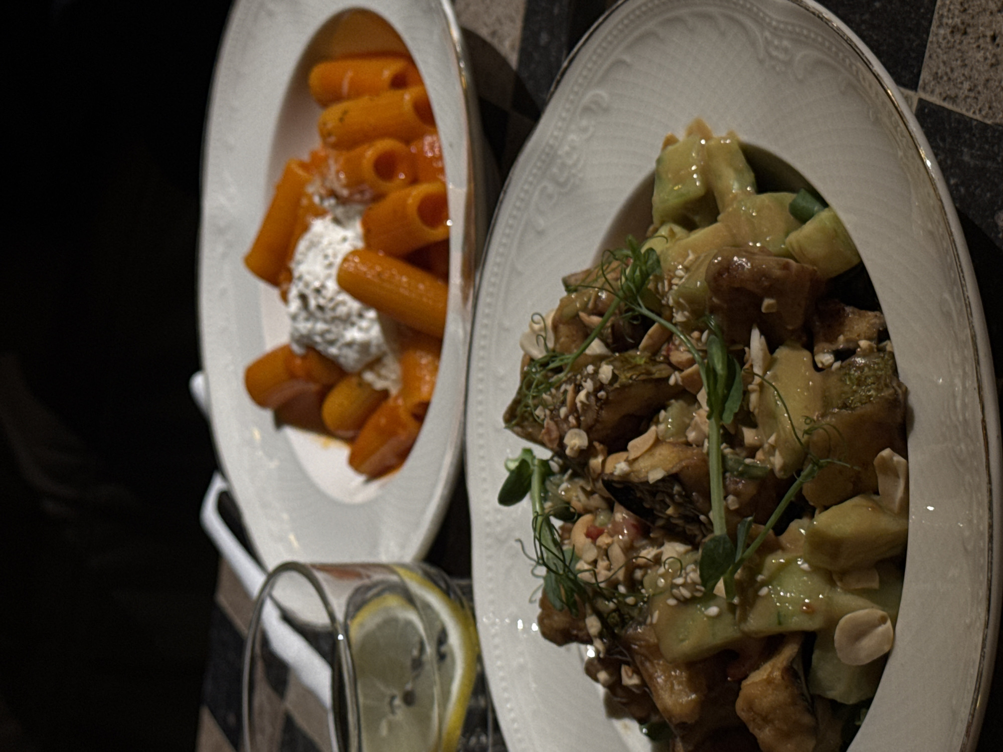 The image shows two plates of food, likely taken indoors. One plate contains pasta with a red sauce and a dollop of creamy white cheese, while the other contains a salad with what appears to be cucumbers, eggplant, and a scattering of seeds or nuts. A glass with a lemon wedge sits near the plates. The setting is not identifiable as Yerevan, Armenia, from visual cues alone.