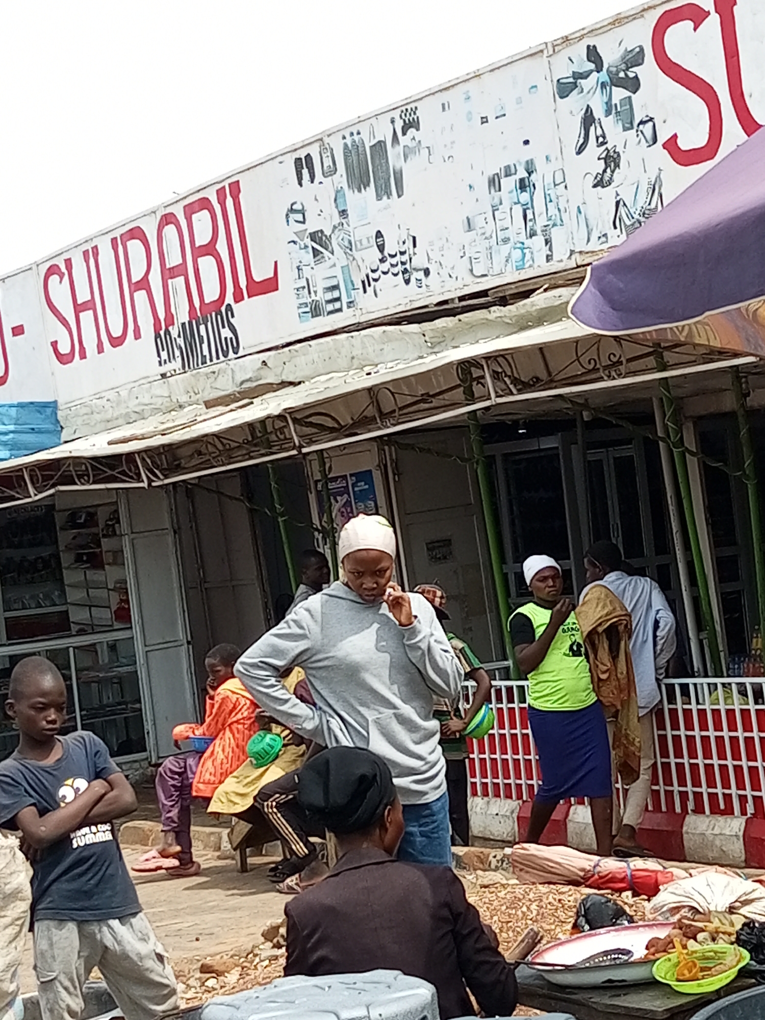 The image depicts a street scene in the city of Gombe, Nigeria. A storefront with a prominent sign reading "SHURABIL COSMETICS" is visible, featuring various illustrations of cosmetic products and shoes. In front of the store, several individuals are present. A young boy stands with his arms crossed, wearing a dark blue t-shirt and gray pants. To his right, a woman in a light gray hooded sweatshirt and a light pink headscarf is holding something to her ear, possibly a phone. Behind her, a child in an orange and yellow outfit is seated. Another person, wearing a bright green t-shirt and a blue skirt, stands with a folded cloth over their shoulder. They appear to be engaged in a conversation with another individual partially visible to the right. The ground in front of the store is littered with various items, including what appears to be dried goods, fabrics, and trays containing food. A low, white fence with red and white stripes runs along the front of the storefront, and a purple awning extends over a section of the sidewalk. The overall impression is that of a busy marketplace or commercial area.