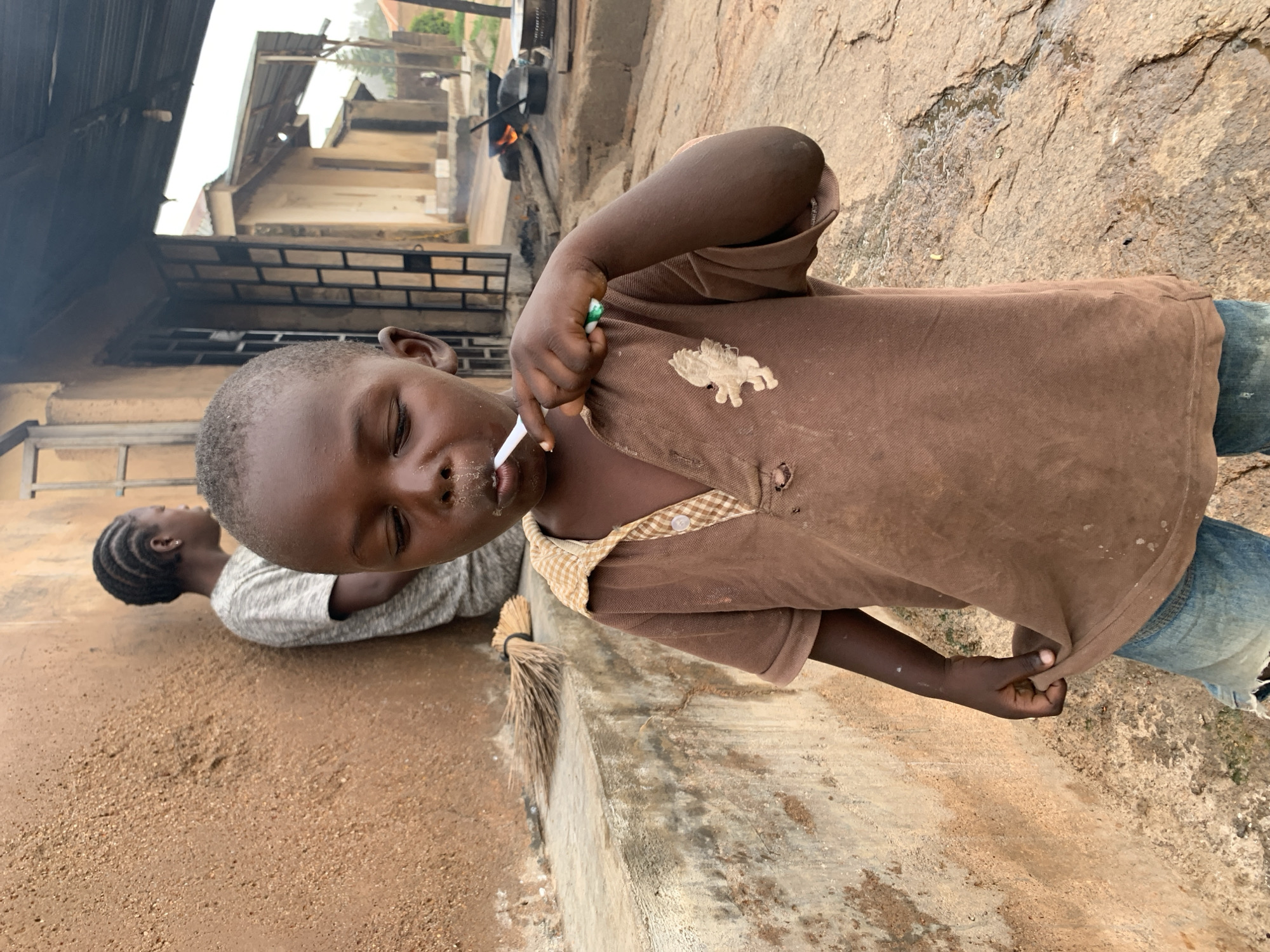 A young boy is brushing his teeth with a white toothbrush outside a building in Jos, Nigeria. He is wearing a brown polo shirt and blue jeans. Behind him, another person, possibly a girl, stands leaning against a textured brown wall. In the background, a cooking fire is visible with pots and pans. The environment appears to be a rural or semi-urban area with dirt ground and simple structures.