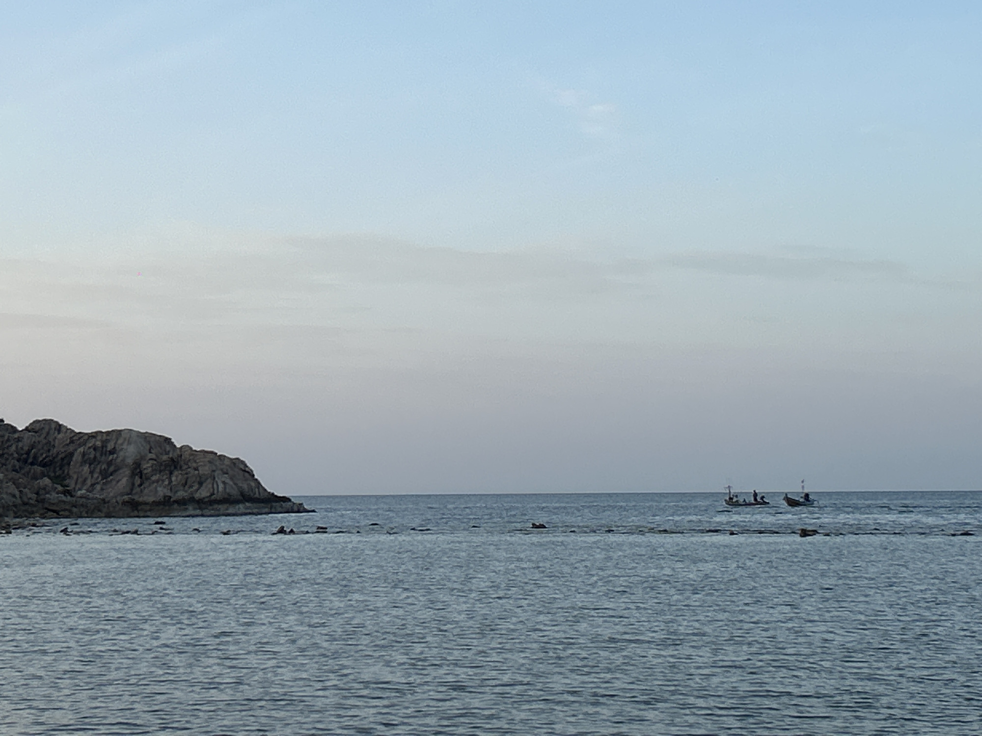 The image shows a serene seascape off the coast of Ko Pha-ngan District, Thailand. In the foreground, the calm, rippling blue sea stretches towards the horizon. A rocky outcrop with dark, textured rocks is visible on the left side of the frame, jutting out into the water. Scattered smaller rocks and submerged formations break the surface of the water along the shoreline. In the distance, two small fishing boats are on the water, each with a few figures onboard. The sky above is a soft gradient of pale blue at the top, transitioning to a hazy, lighter hue near the horizon, suggesting either dawn or dusk. The overall atmosphere is peaceful and tranquil.