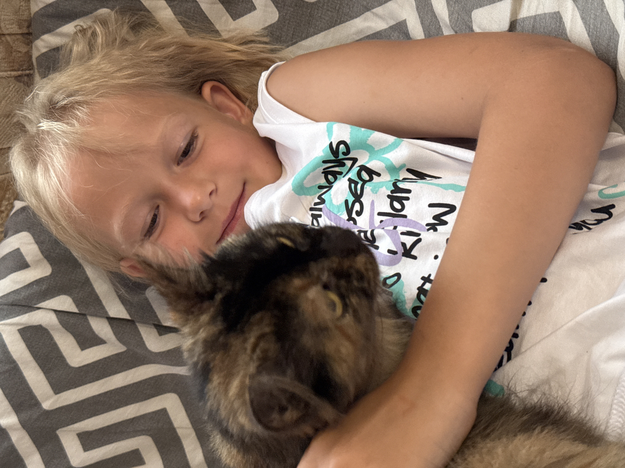 A young girl with blonde hair is lying down and hugging a tortoiseshell cat. The girl is wearing a white t-shirt with text and graphics. The cat is nestled into the girl's chest and is partially obscuring her. They are lying on a surface with a striped pattern. The image appears to be taken indoors, likely in a home setting. The location in Sovietskyi, Ukraine is not determinable from the image.