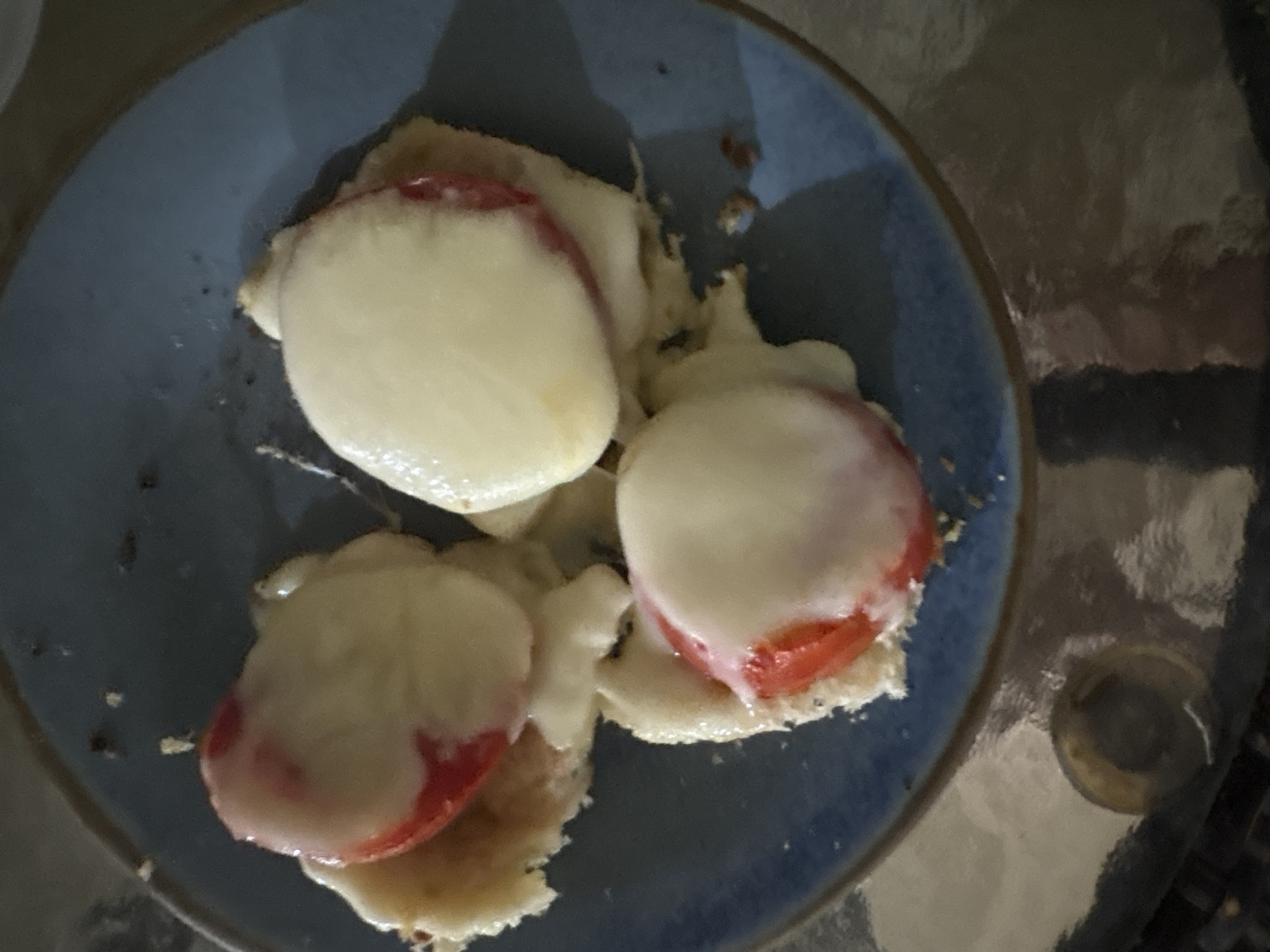 A close-up, top-down view shows three small, open-faced sandwiches on a blue plate. Each sandwich consists of a round piece of bread topped with a slice of tomato and melted white cheese. The sandwiches are arranged in a triangular formation. The plate is dark blue and has a slightly reflective surface. The background is dark and out of focus. The image appears to be taken in a dimly lit indoor setting, possibly a restaurant or home kitchen. There is no information about Ashvem, India visible in the image.