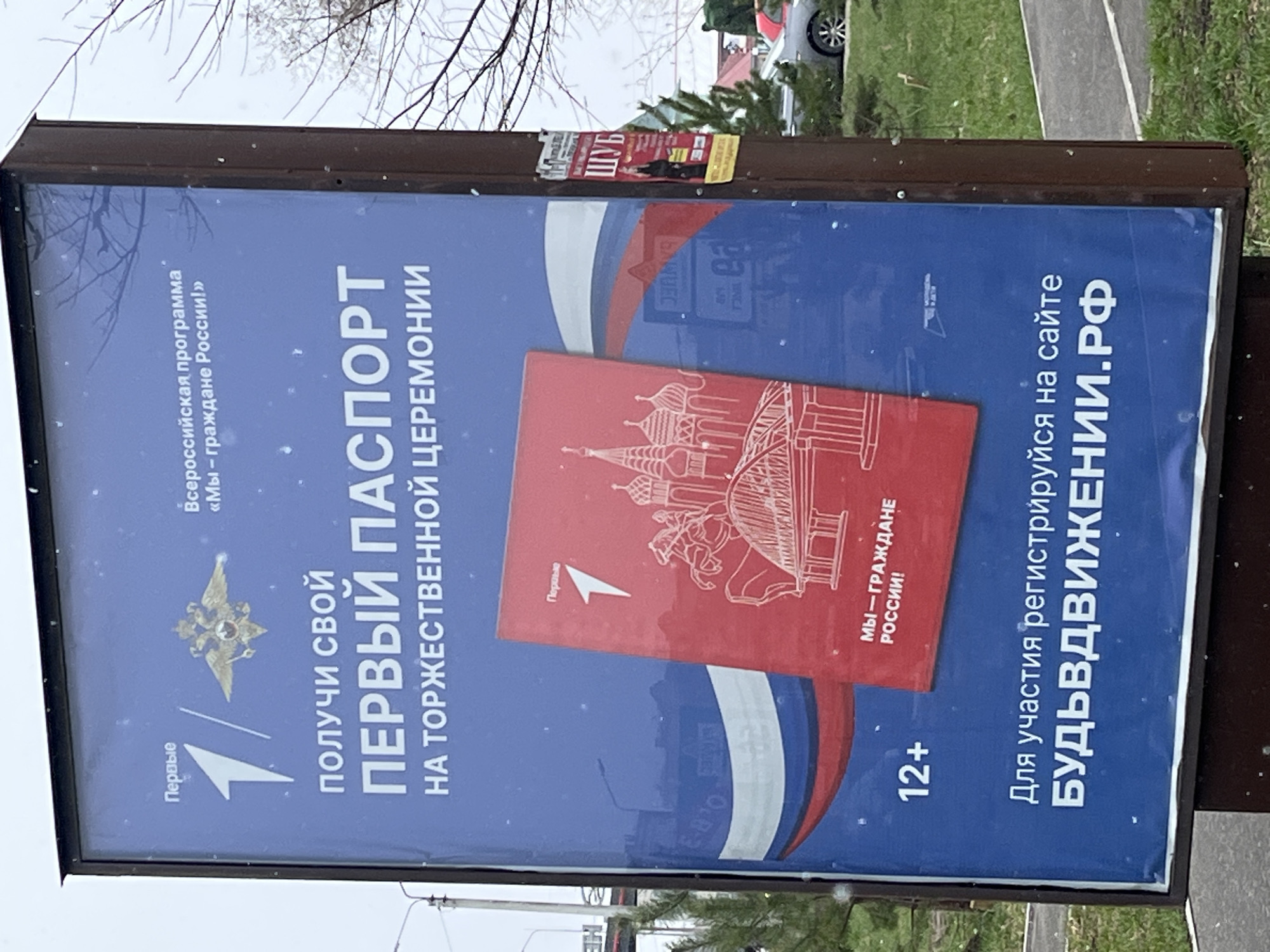 Here's a detailed description of the image:
**Overall Scene:** The image shows an advertisement, likely on a bus stop or roadside display, for a program focused on issuing the first passport to citizens of Russia. The setting appears to be outdoors with some greenery and a road visible in the background.
**Advertisement Details:**
* The advertisement has a blue background with white and red elements.
* The main text includes phrases like "Get Your First Passport" and "At the Solemn Ceremony."
* There is an image of the Russian passport, a red booklet with architectural illustrations.
* The text "We are Citizens of Russia!" is also prominently featured.
* The ad indicates an age range of 12+ and includes a website address, "БУДЬВДВИЖЕНИИ.РФ", for registration.
**Additional Details:**
* There is a "Первые" (First) label and an image of the Russian coat of arms.
* A small poster labeled "ШУБ" is taped to the top of the advertisement frame.
* The overall condition suggests the advertisement has been in place for some time, possibly with some weathering or wear.
**Location:** The image has been labeled to be in Chaikovsky, Russia, though there's no specific feature within the advertisement or visible in the surroundings that explicitly confirms that location.