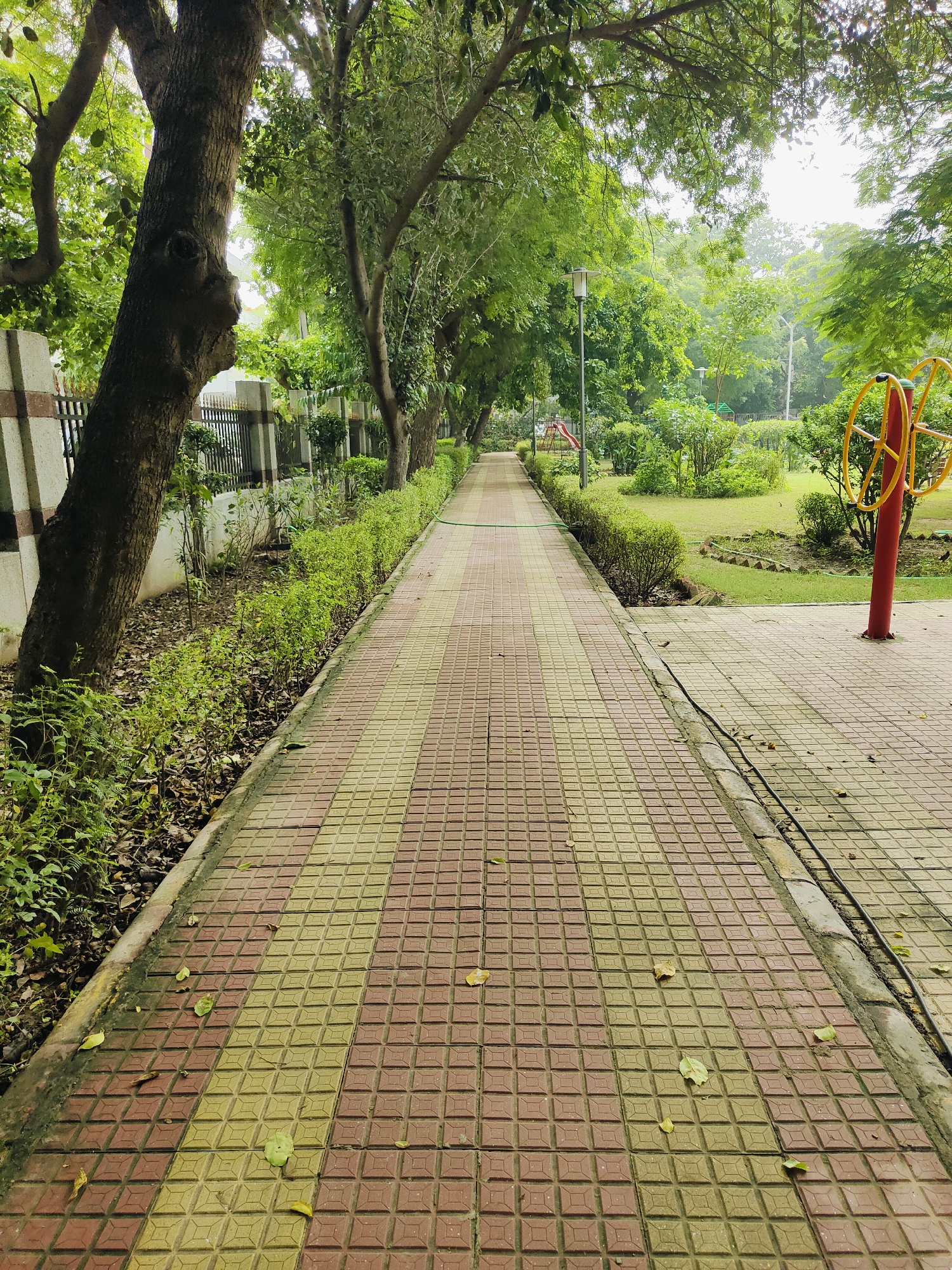 The image depicts a paved walkway in a park in Gurgaon, India. The walkway is made of square tiles in alternating shades of red and yellow, and it stretches into the distance, flanked by lush greenery and trees. On the left side of the walkway, there is a low, manicured hedge and behind it, a concrete wall with a black metal fence. Large trees with thick trunks grow along this side, their branches arching over the walkway. On the right side of the walkway, there is a grassy area with more trees, bushes, and what appears to be outdoor exercise equipment. The sky is overcast, contributing to a soft, diffused light. A red and yellow circular structure, part of the exercise equipment, stands prominently on the right. There are no people visible in the image.