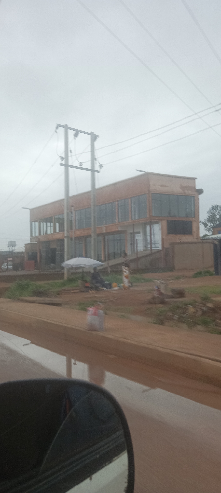 The image depicts a two-story building under construction in Bukuru, Nigeria. The building has a rectangular footprint with large window openings on both floors. The exterior appears to be unfinished. In front of the building, there is an area with some vegetation and loose dirt. An electrical utility pole stands to the left of the building. Near the front of the building, there are people under an umbrella. A bag is partially visible on the side of the road. A side mirror of a vehicle is visible in the lower left corner. The sky is overcast.