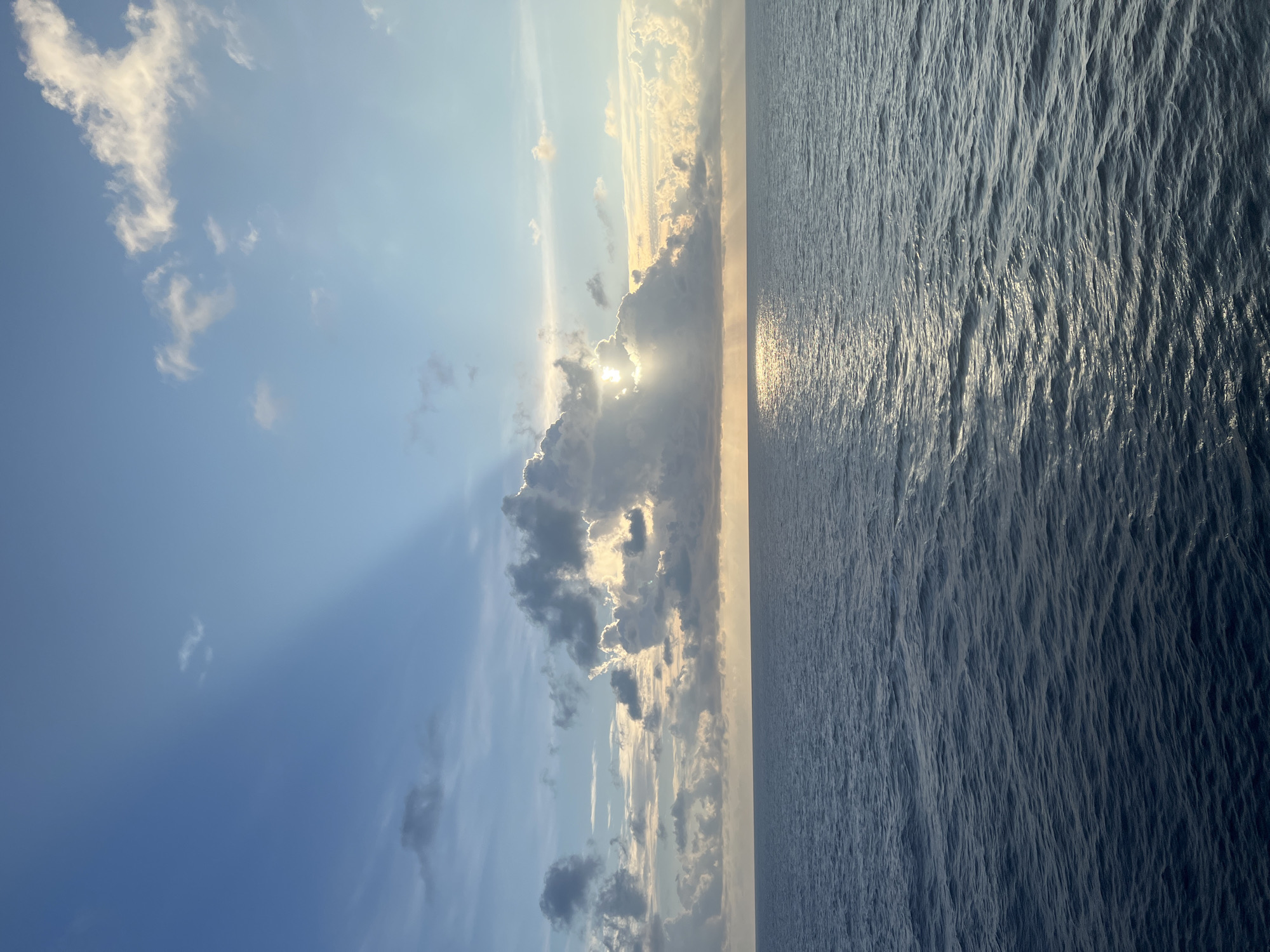 The image depicts a vast expanse of the ocean under a sky with scattered clouds during what appears to be sunrise or sunset. The sun is partially obscured by a large cloud formation in the center of the frame, casting rays of light downwards onto the water's surface. The ocean is a deep blue, with small waves creating ripples and reflecting the sky's colors. The horizon line is clearly defined, separating the water from the sky. The sky is a gradient of blues, with some lighter, warmer tones near the horizon, suggesting the sun's proximity. There are no people, animals, or man-made structures visible in the image, making it a natural landscape scene. The location is stated to be Unknown, Unknown.