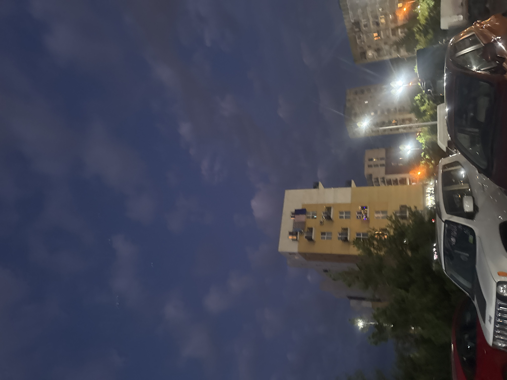 In Tbilisi, Georgia, at dusk or night, the sky is a deep blue with scattered clouds. Several apartment buildings of varying heights are visible, some illuminated with lights. Laundry is hanging on balconies of one building. The foreground shows parked cars, including a white Jeep with a black hood decal and a red car. Streetlights are on, casting light and creating lens flares. Trees are also present in the scene. The overall impression is that of an urban residential area at night.