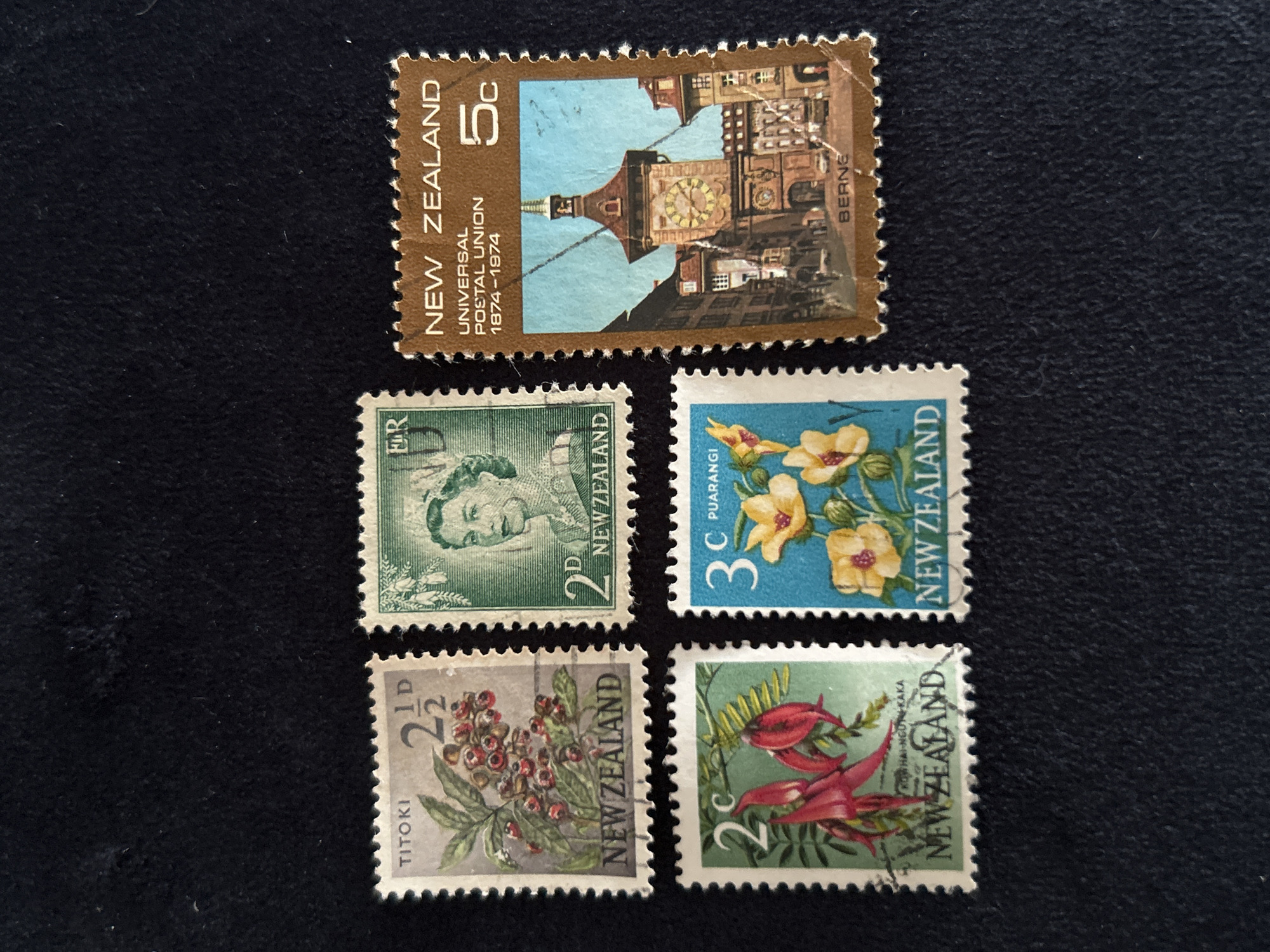 Five New Zealand postage stamps are displayed against a dark background. The stamps are arranged in two rows, with three stamps in the top row and two in the bottom row.
The top left stamp depicts a branch with red berries and green leaves, labeled "TITOKI" and "NEW ZEALAND". It has a denomination of "2 1/2 D".
The top middle stamp features a portrait of Queen Elizabeth II, with "ER" and "NEW ZEALAND" visible. Its denomination is "2 D".
The top right stamp is brown and features an image of a clock tower in a European city, with the text "NEW ZEALAND", "UNIVERSAL POSTAL UNION 1874-1974", and "5c". The clock tower appears to be in Bern, Switzerland, as indicated by the label "BERNE" at the bottom of the stamp.
The bottom left stamp displays a close-up of two red flowers with green leaves, labeled "NEW ZEALAND". It has a denomination of "2c". Below the flowers, partially obscured, is text that appears to read "KOHAI-NGUTU-RAKA".
The bottom right stamp features yellow flowers with green leaves on a blue background, labeled "NEW ZEALAND". It has a denomination of "3c" and "PUARANGI".