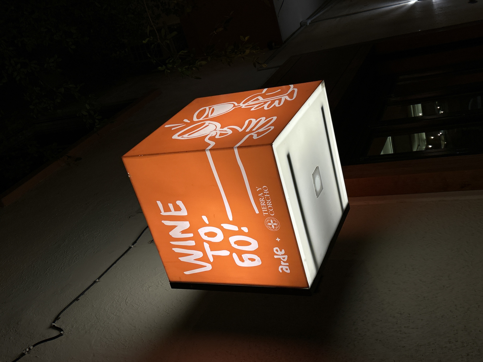 A brightly lit, orange, cube-shaped sign hanging on the wall of a building. The sign reads "Wine To-Go!" in white lettering. Above that are white line drawings of two glasses, one filled with liquid, and two hands reaching up. The other visible side of the cube sign is white with "arde + TIERRA Y CORCHO" printed in smaller letters. The wall is a rough, light gray texture with some electrical wires running up it. Above the sign, a dark window is visible. The sign appears to be advertising a wine service in a location, and based on the brand, is likely Mazatlán, Mexico. The scene is dimly lit, implying it is nighttime.