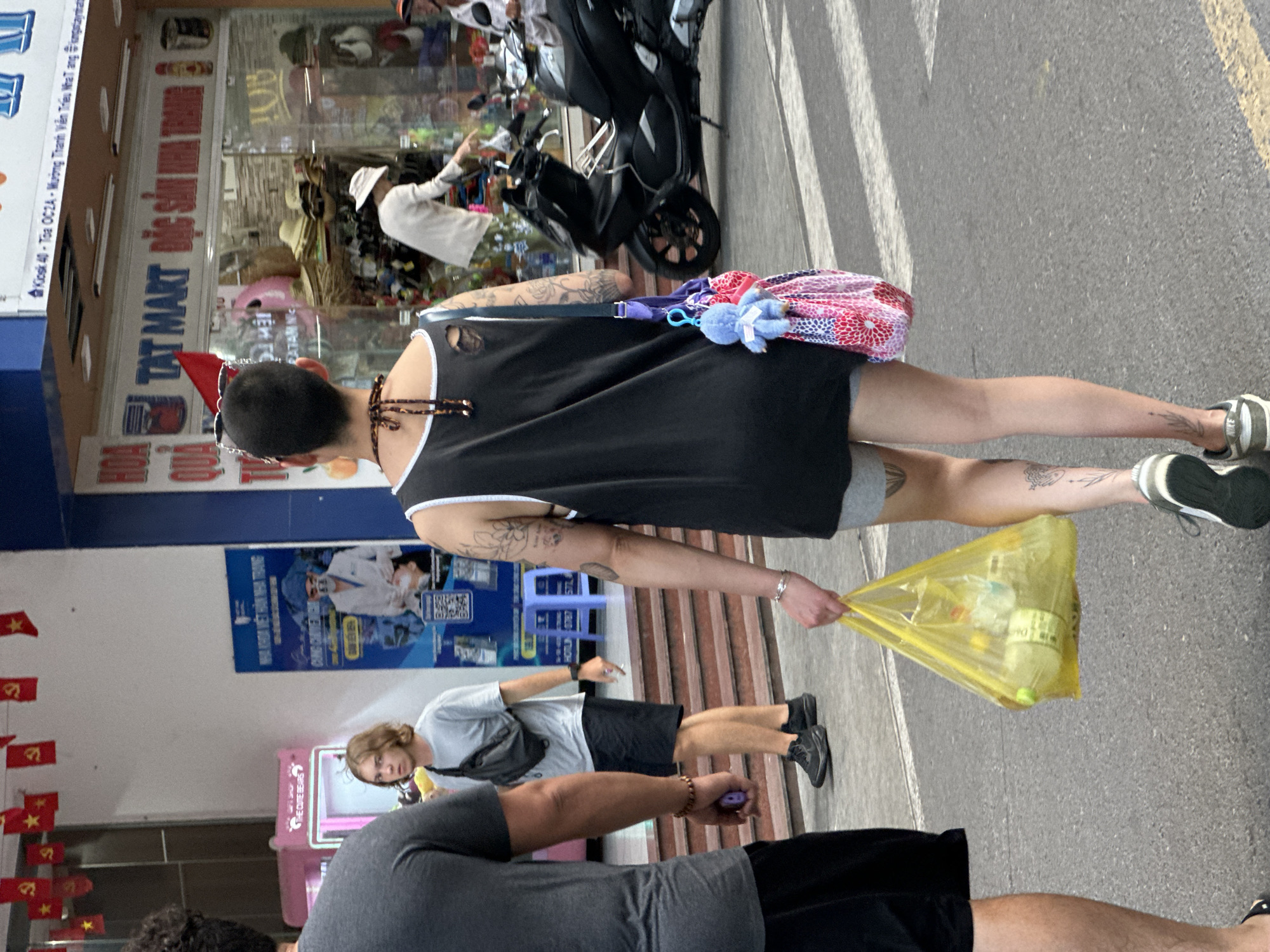 The image depicts a street scene, appearing to be in the city of Bac Nha Trang Ward, Vietnam, with multiple people visible. In the immediate foreground, two individuals are partially visible, both wearing dark clothing. Slightly further back, a third person wearing a light-colored t-shirt and dark shorts is present. A fourth person, walking on the street, wears a black tank top, grey shorts, and athletic shoes; they are carrying a yellow plastic bag.
In the background, a storefront is visible. The storefront signage reads "TAT MART", and other text is present in Vietnamese. Further in the background, a person wearing a white shirt and hat appears to be inside the storefront. Additionally, parked motorbikes are noticeable in the background. Red flags decorate the foreground, near the storefront.