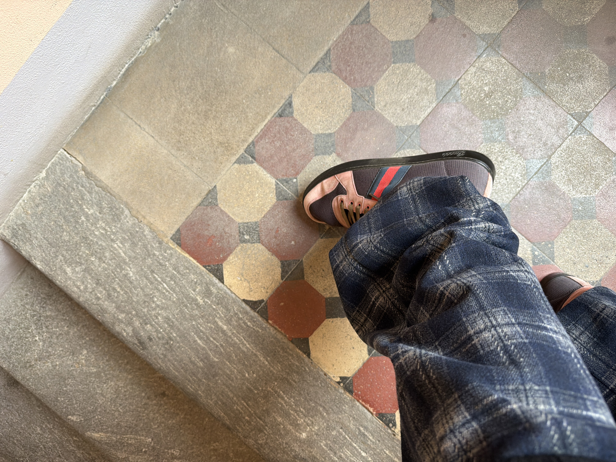The image shows a person's-eye view of a descent on a staircase. The person is wearing blue plaid pants and a sneaker with pink, brown, and navy blue details. The staircase is stone and the floor is tiled in an octagonal pattern in shades of red, tan, and gray. It's plausible that this is in Milan, Italy, due to the architectural style of the tiled floor and stone staircase.