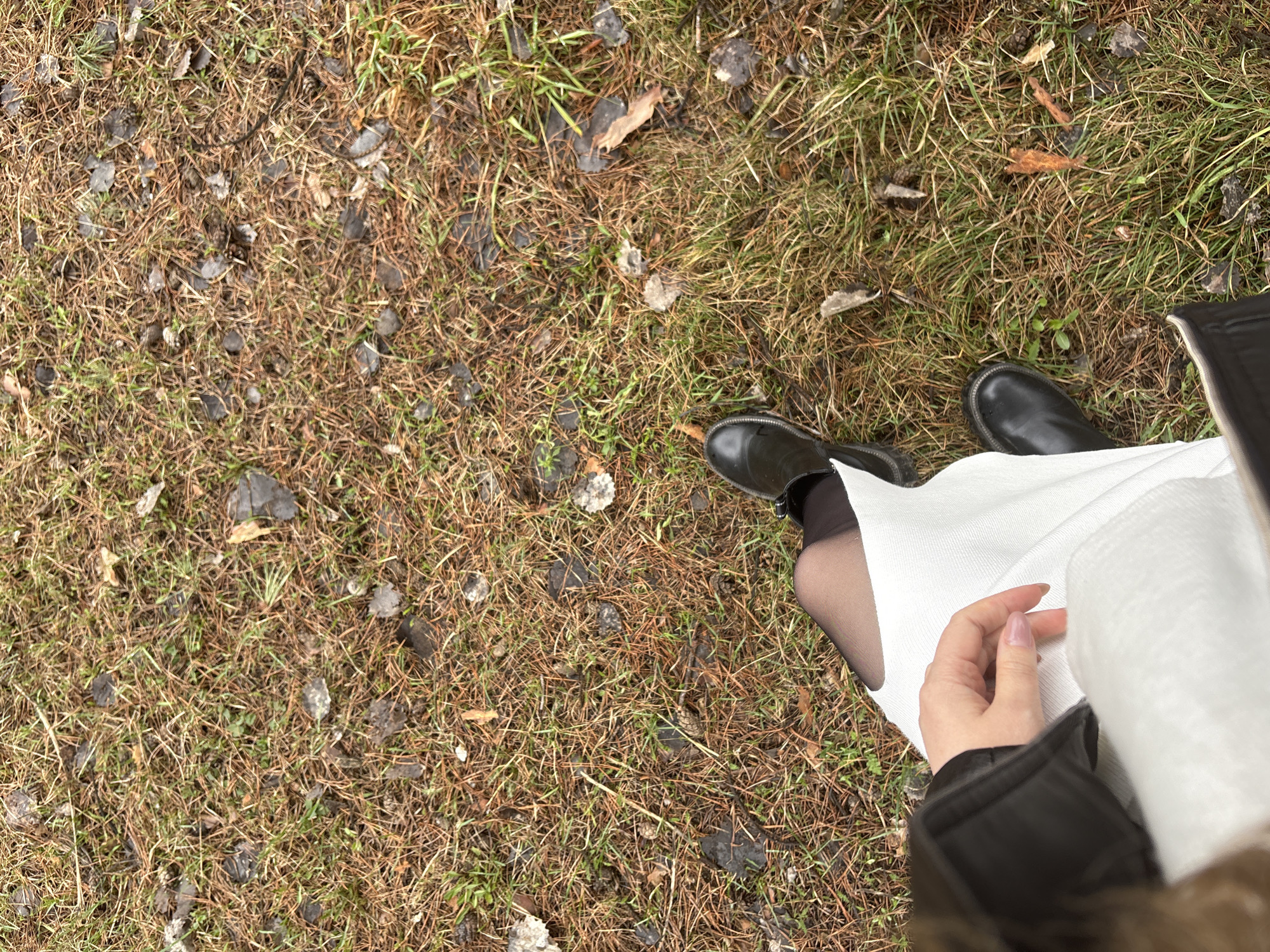 An overhead shot depicts a person standing on a ground surface composed of mixed dry grass, green grass, small leaves, and dark-colored debris. The person is wearing black boots and black stockings, along with a white dress or cloth draped over their lap. The person's hand, with visible fingernails, is also visible on the white fabric. The edge of a black jacket is visible in the lower right corner. There are no clear indicators of the location being Chaikovsky, Russia. The environment is dominated by the ground surface and the person's attire, suggesting a focus on personal appearance within a natural setting.