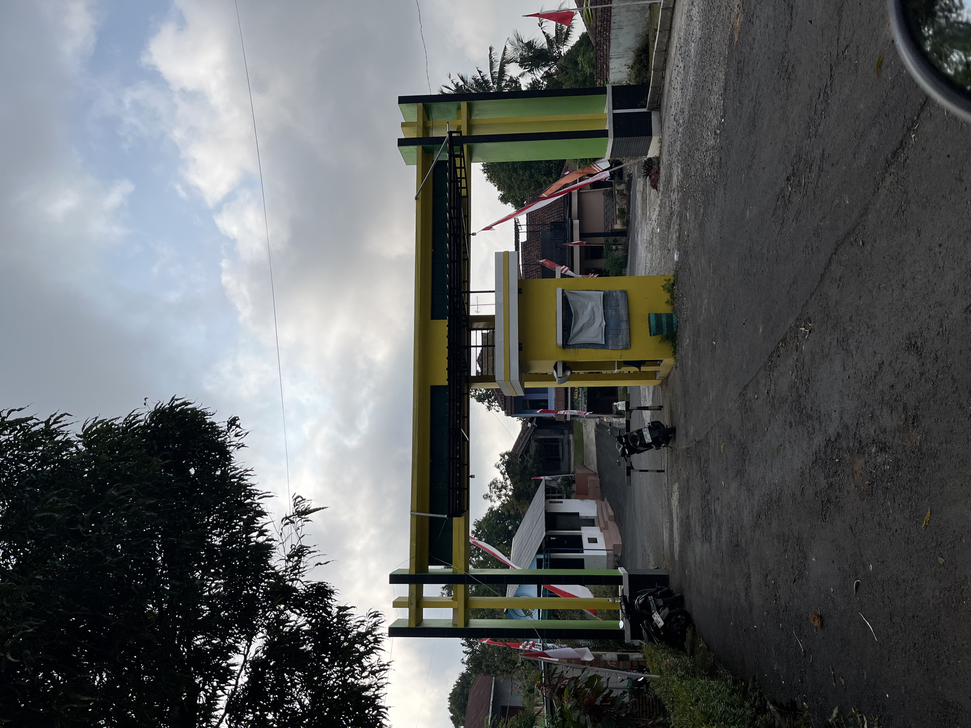 The image depicts an outdoor scene under a cloudy sky. In the foreground, a dark asphalt road leads towards a large, yellow and green archway. The archway has vertical black and green pillars supporting a horizontal yellow beam, adorned with black decorative elements. To the left and right of the archway, there are structures that appear to be part of an entrance or administrative building, painted in yellow and white. Several Indonesian flags, red and white, are displayed on poles along the entrance.

Parked on the road are two motorcycles to the left of the archway and one motorcycle to the right. Beyond the archway, a paved road continues, with several buildings visible in the background, including houses with red tile roofs and some with white walls. Lush green trees and foliage surround the area, suggesting a semi-rural or suburban environment. The overall atmosphere is calm and overcast. The image captures an entrance to a complex or community in Gembong, Indonesia, likely a place with a local administration or a residential area.