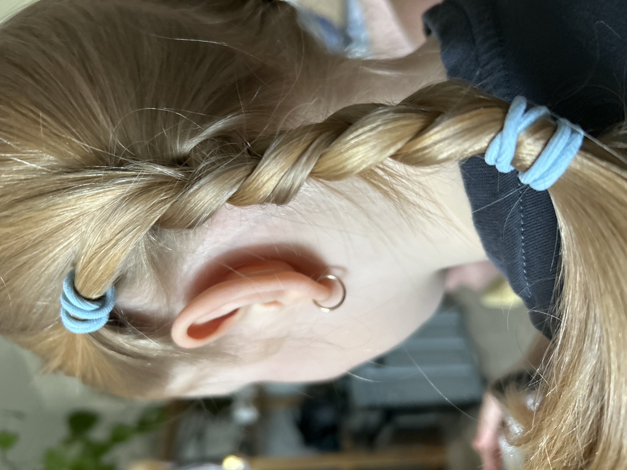 Close-up shot of a fair-skinned person with blonde hair styled in a braid, secured with light blue hair ties. The person is wearing a small gold hoop earring in their left ear. A portion of a dark-colored garment is visible near the braid. The background is blurred. The location is not specified as Chaikovsky, Russia, in the image.