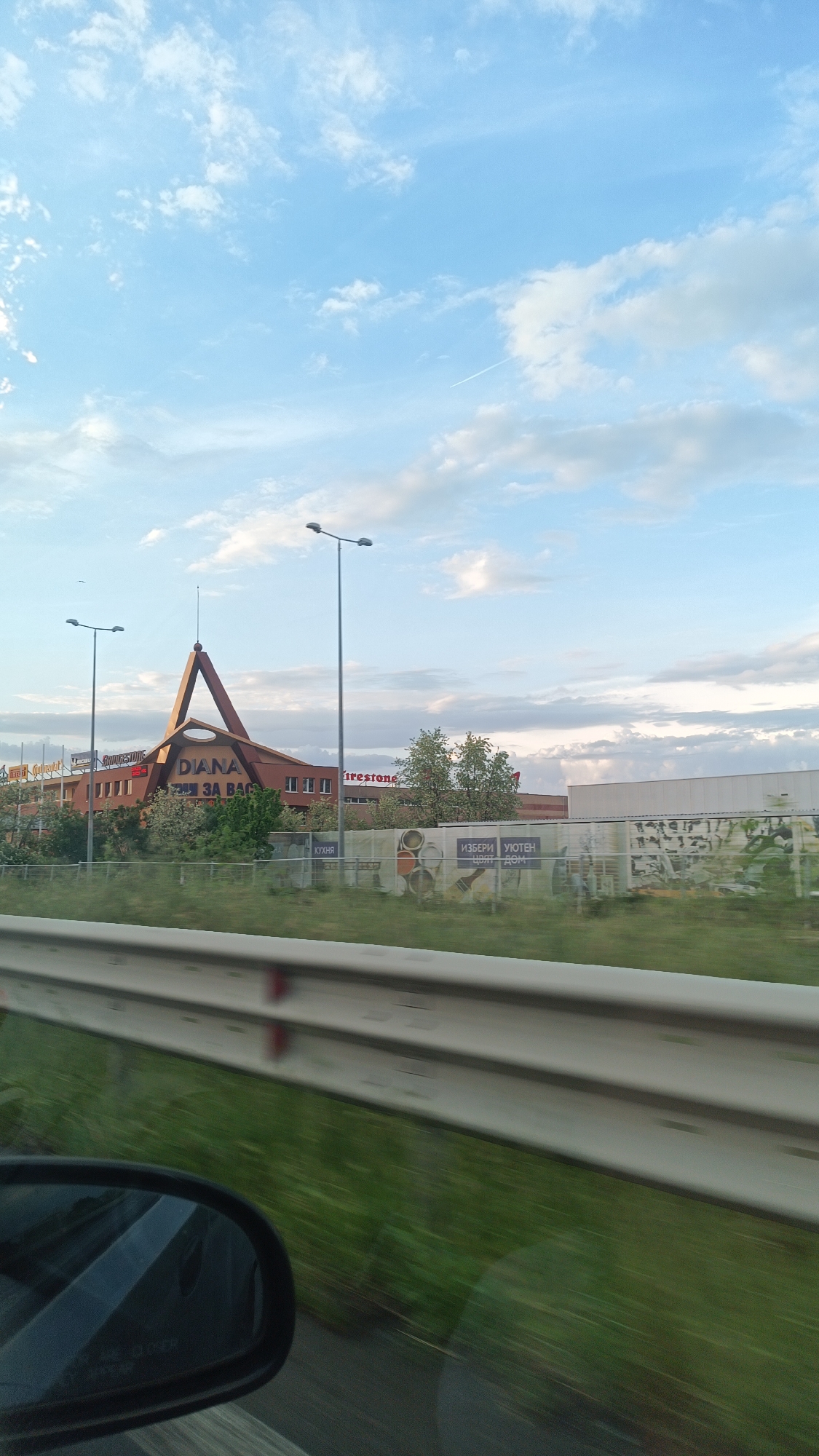 A slightly blurry, medium-shot photograph taken from a moving vehicle captures the Diana Shopping Center in Burgas, Bulgaria, along with the surrounding environment. The shopping center, a long, low, red-brick building, features a distinct triangular arch above the entrance. The name "DIANA" is prominently displayed along with other signage in Cyrillic. Brand names like "Bridgestone" and "Firestone" are also visible on the building's facade.
In front of the building, a grassy area is bordered by a temporary construction fence. The fence is adorned with large advertisements. Streetlights are positioned in front of the building.
The foreground includes a grey guardrail and blurred green vegetation, indicating motion. Part of the car's side mirror is visible in the lower left corner.
Above the scene, the sky is blue with scattered white clouds.