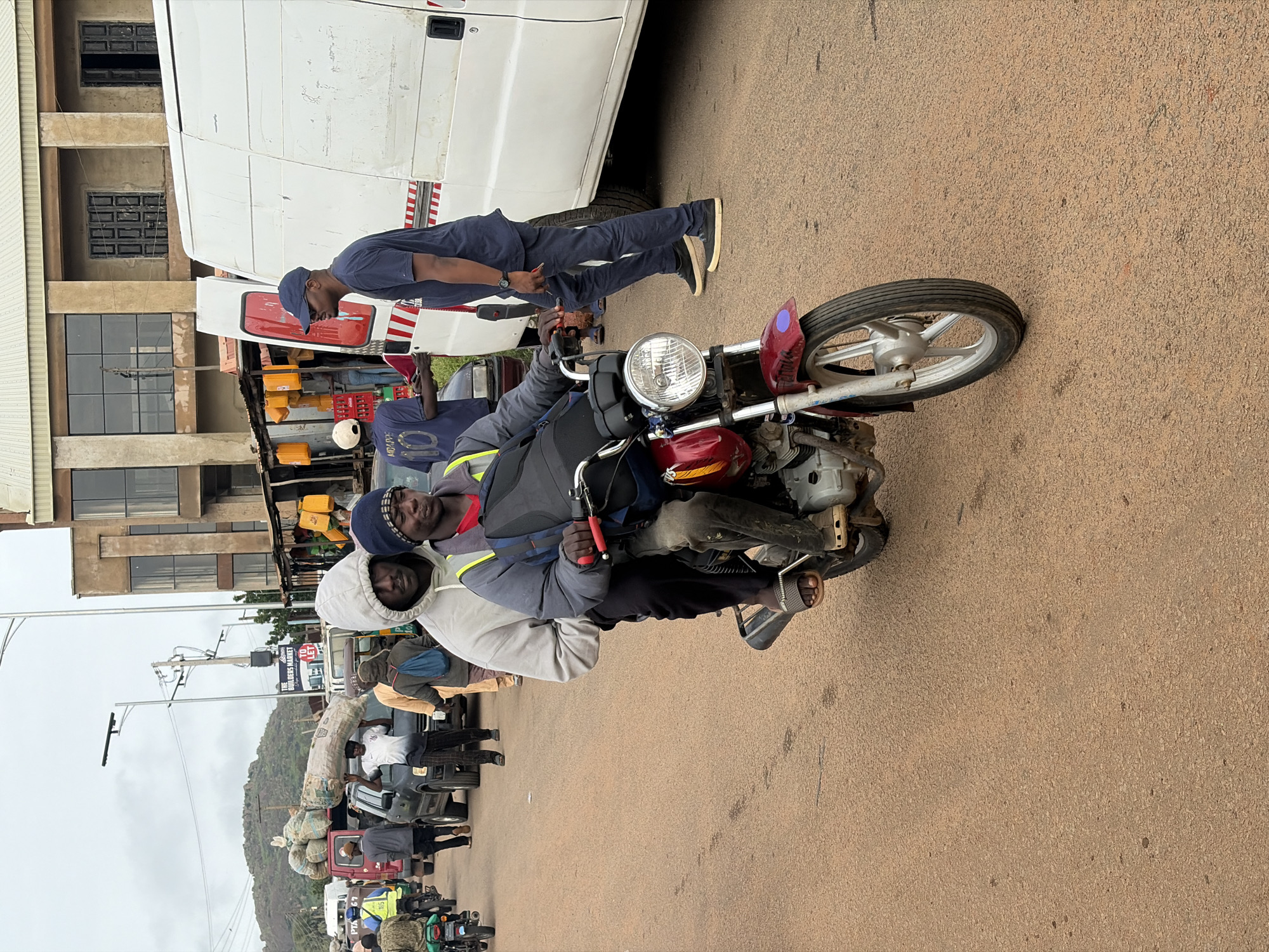 The image captures a busy street scene in Gura Top, Nigeria. In the foreground, two men are riding a red motorcycle. The driver wears a blue jacket, grey hoodie, and a beanie, while the passenger in front has a grey hoodie and a blue backpack. They are positioned on a dusty, reddish-brown paved road, with other vehicles and pedestrians visible in the background. To the right, a large white van is parked, and a man in a dark blue t-shirt and cap stands near it. Further back, several cars, motorcycles, and people carrying goods are present. Buildings with muted tones and windows line the side of the road, suggesting a commercial or residential area. The sky is overcast. The overall impression is one of active daily life and commerce on a Nigerian street.