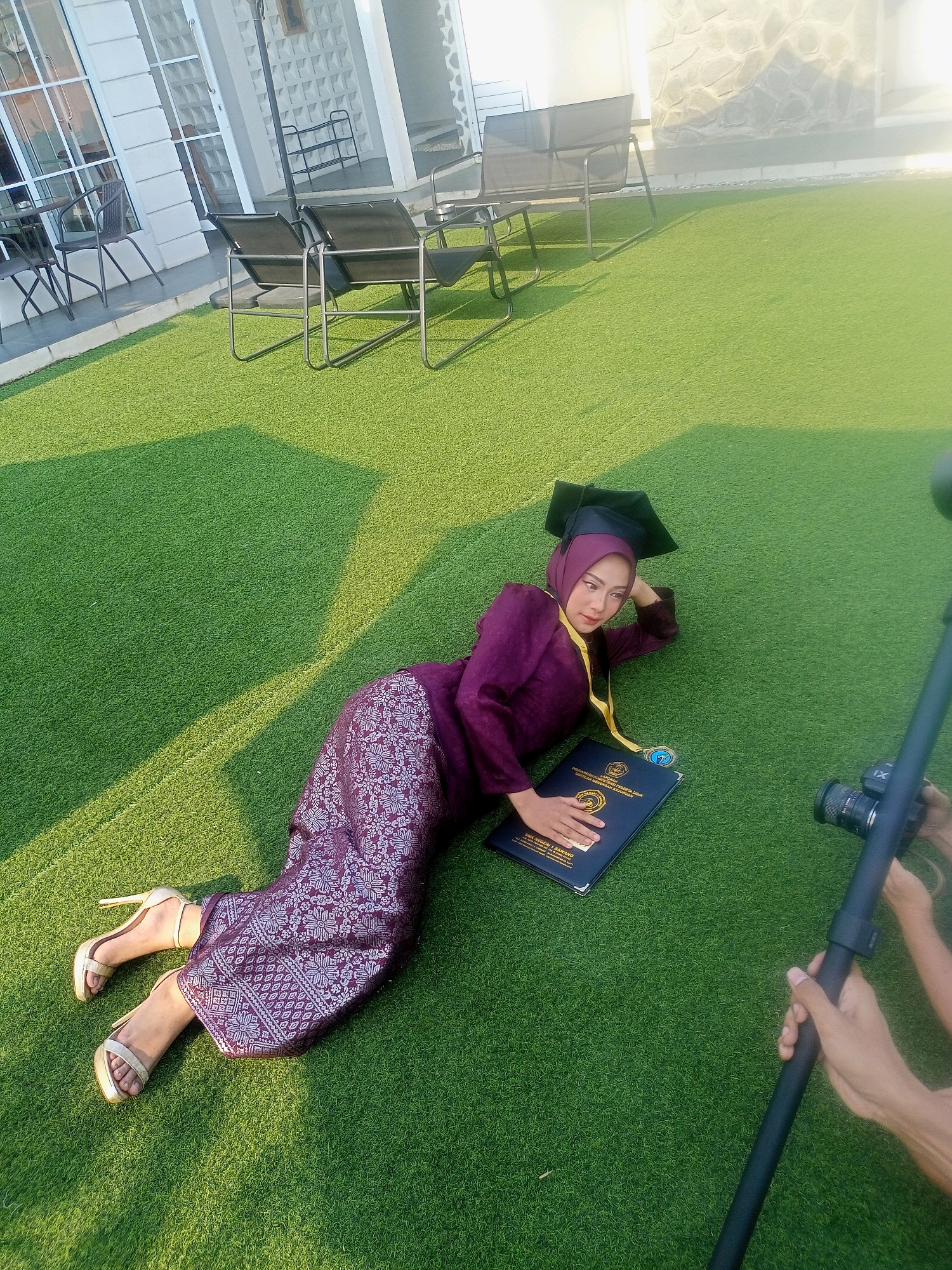 The image depicts a young woman lying on artificial grass, dressed in a graduation gown and cap. She is wearing a purple hijab and a patterned purple skirt with gold high heels. In front of her lies a dark blue folder, likely containing her graduation documents, with a medal draped around her neck. To the right of the frame, a camera on a tripod is visible, with hands holding it, suggesting a photoshoot is in progress. The background features outdoor furniture and a building with modern architecture, consistent with a location in Blambangan, Indonesia. The scene is illuminated by natural sunlight, casting shadows on the grass.