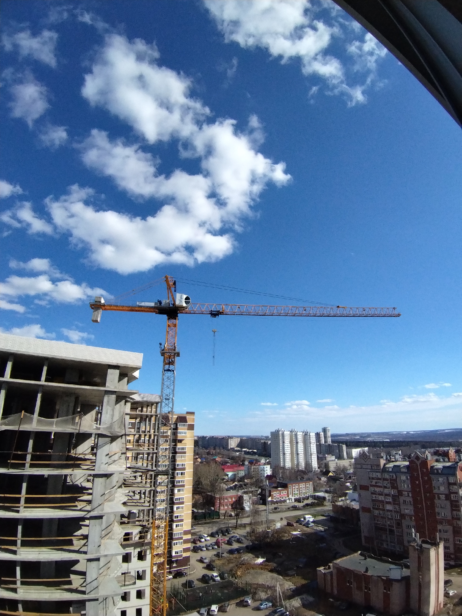 The image depicts a construction site in Izhevsk, Russia, under a bright blue sky with scattered clouds. A tall construction crane, painted orange and gray, dominates the scene, positioned alongside a partially constructed, multi-story building. The building's concrete skeleton is visible, with open spaces where windows and walls will eventually be.
Beyond the construction site, the cityscape of Izhevsk stretches into the distance. Varied architectural styles are apparent in the buildings, ranging from older, Soviet-era structures to more modern high-rises. Cars are parked and moving on the streets below. In the upper right of the frame, part of a building intrudes into the shot.
