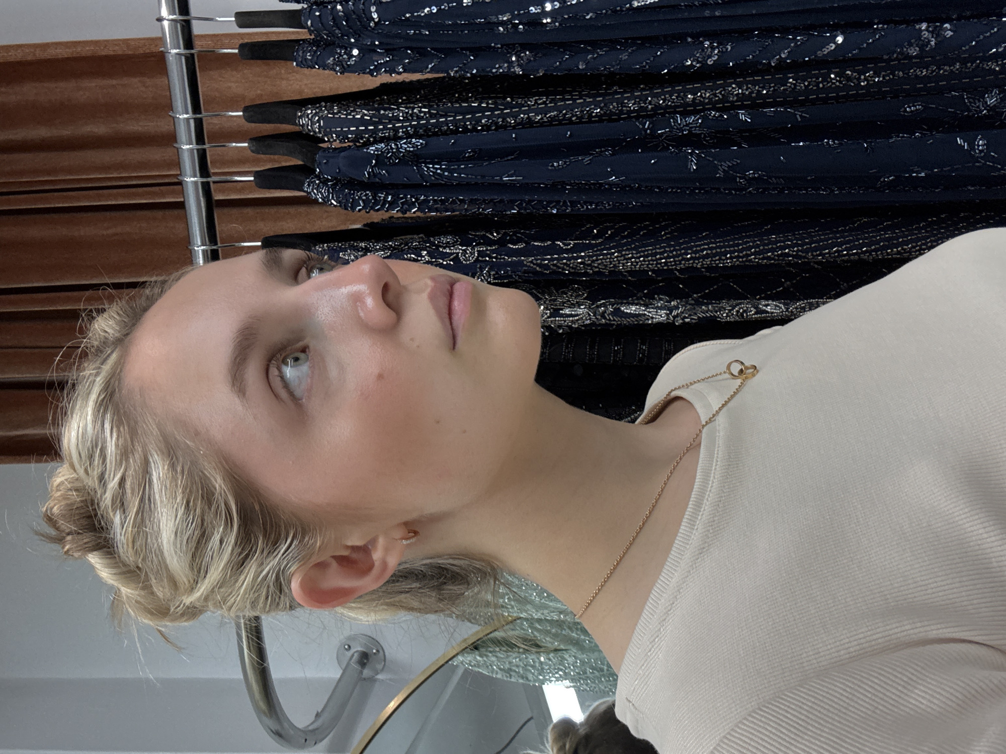 A young Caucasian woman with blonde hair pulled into a messy bun is looking to her right. She is wearing a cream-colored ribbed long-sleeve shirt and a delicate gold necklace with a circular pendant. Her left ear, adorned with a small hoop earring, is visible. Behind her, a rack of dark blue, heavily embellished dresses hangs against a brown velvet curtain. The background also shows a sliver of a silver railing and a mirrored surface. The lighting suggests an indoor setting, possibly a boutique or dressing room.