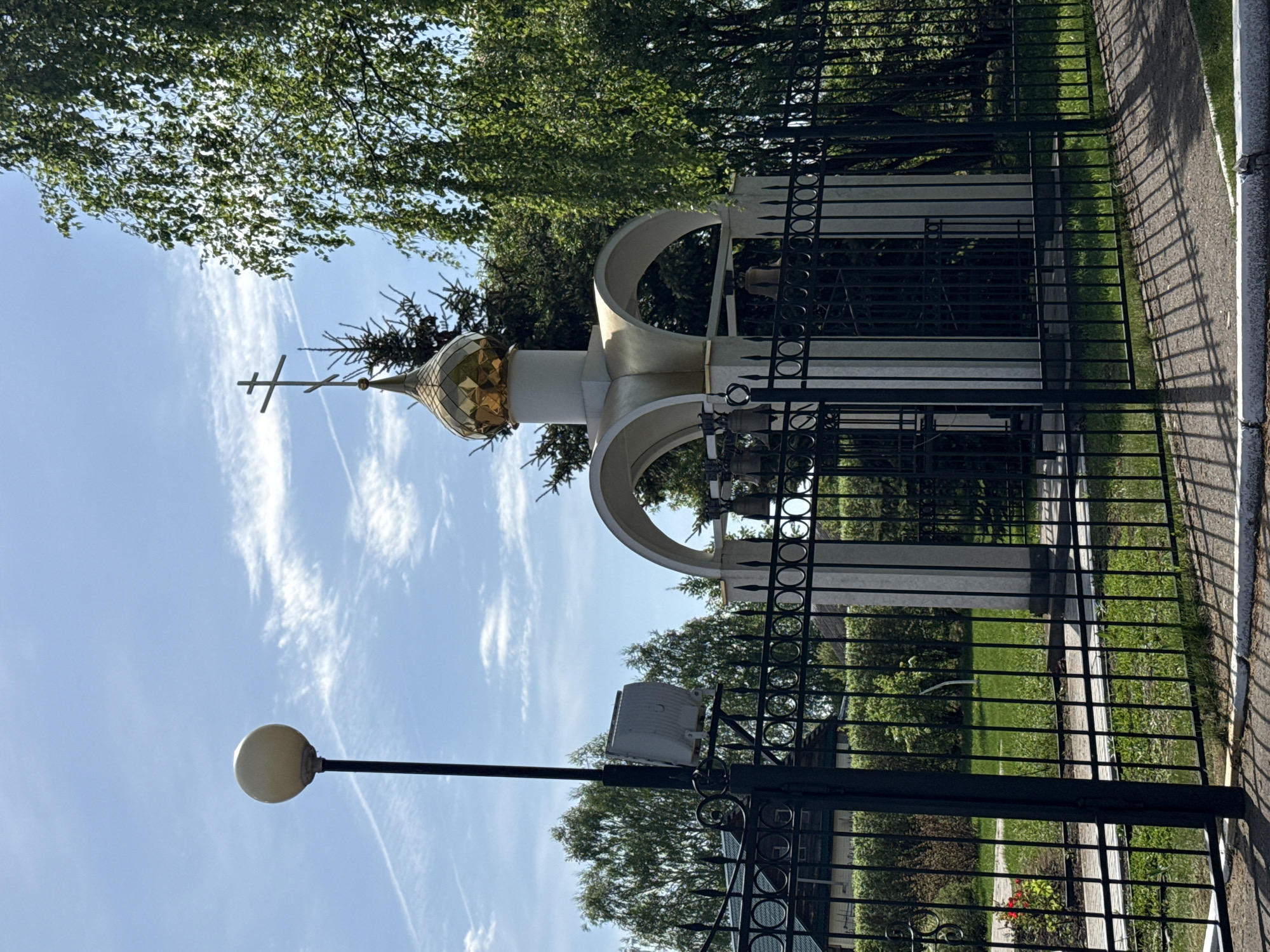 The image depicts a monument in Votkinsk, Russia, likely a memorial for an Orthodox church. The monument has a gold-colored, onion-shaped dome topped with a cross. The dome is supported by white arches and pillars. A black, wrought-iron fence surrounds the monument. A lamppost stands to the left. Trees are visible in the background, along with a blue sky dotted with clouds. A sidewalk runs along the right side of the fence.