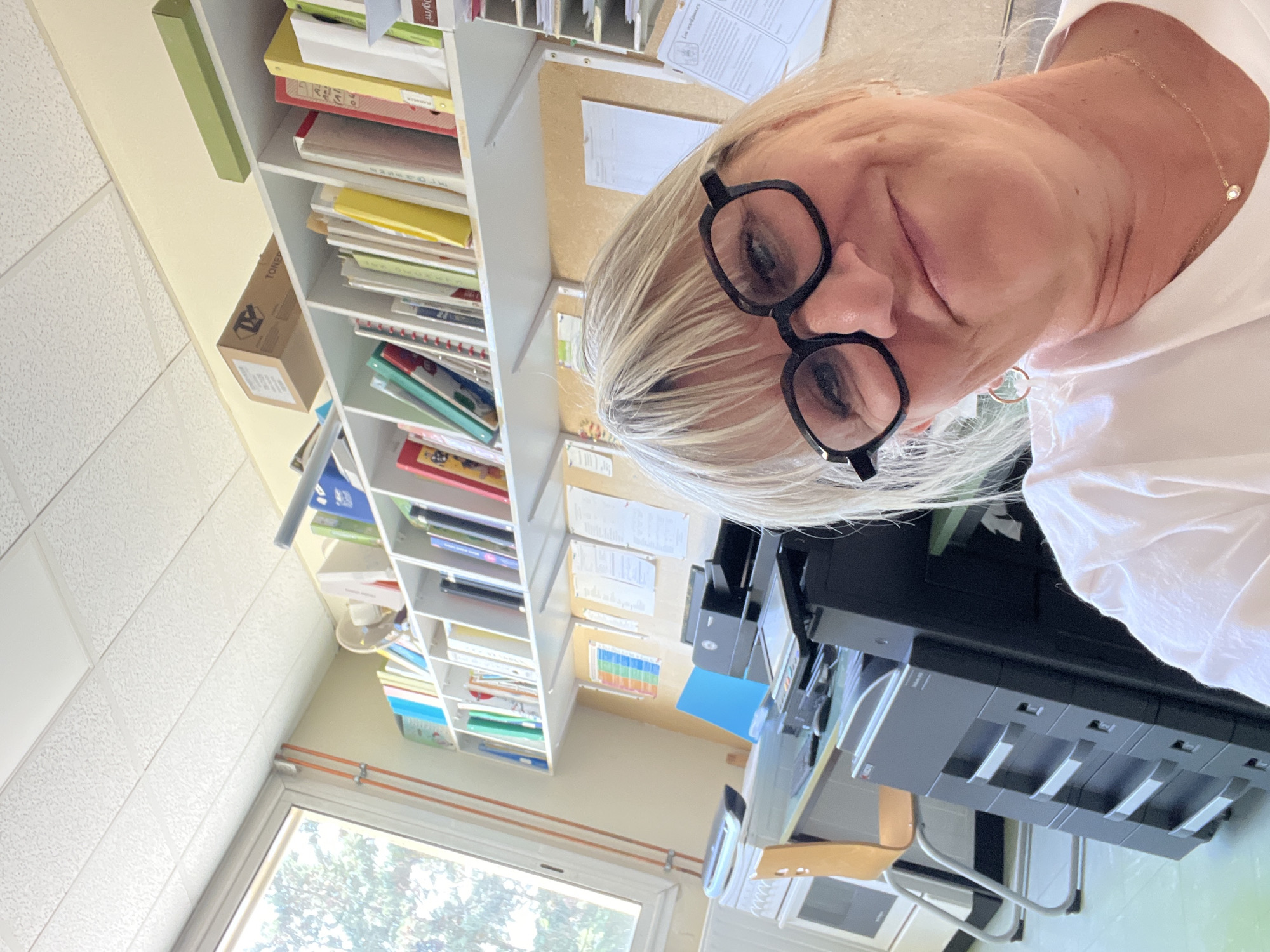 The image shows a woman with blonde hair and glasses taking a selfie in what appears to be an office or classroom environment. She is wearing a white t-shirt and a necklace. Behind her, there are shelves filled with books and binders, a printer, a desk, and a window. The room has a white drop ceiling. The overall setting suggests an administrative or educational workspace. The location in the city of La Fouillouse, France, is not explicitly evident from the image itself.