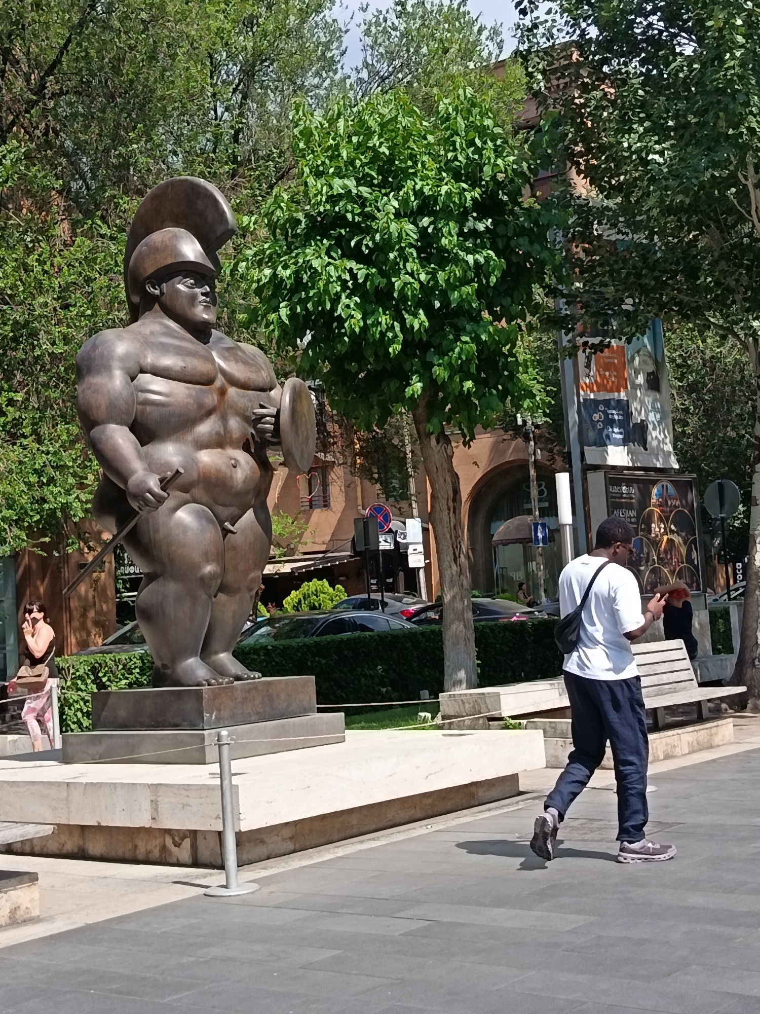 A bronze statue of a muscular, heavyset gladiator stands on a stone pedestal in a city park. The gladiator is wearing a helmet and holding a shield and sword. A man in a white t-shirt and dark pants walks away from the camera in the foreground, looking at his phone. A woman in a pink and white striped outfit is visible to the left of the statue, partially obscured by foliage. The background features trees, buildings, and posters on a pole. The scene is lit by sunlight, casting shadows on the ground.