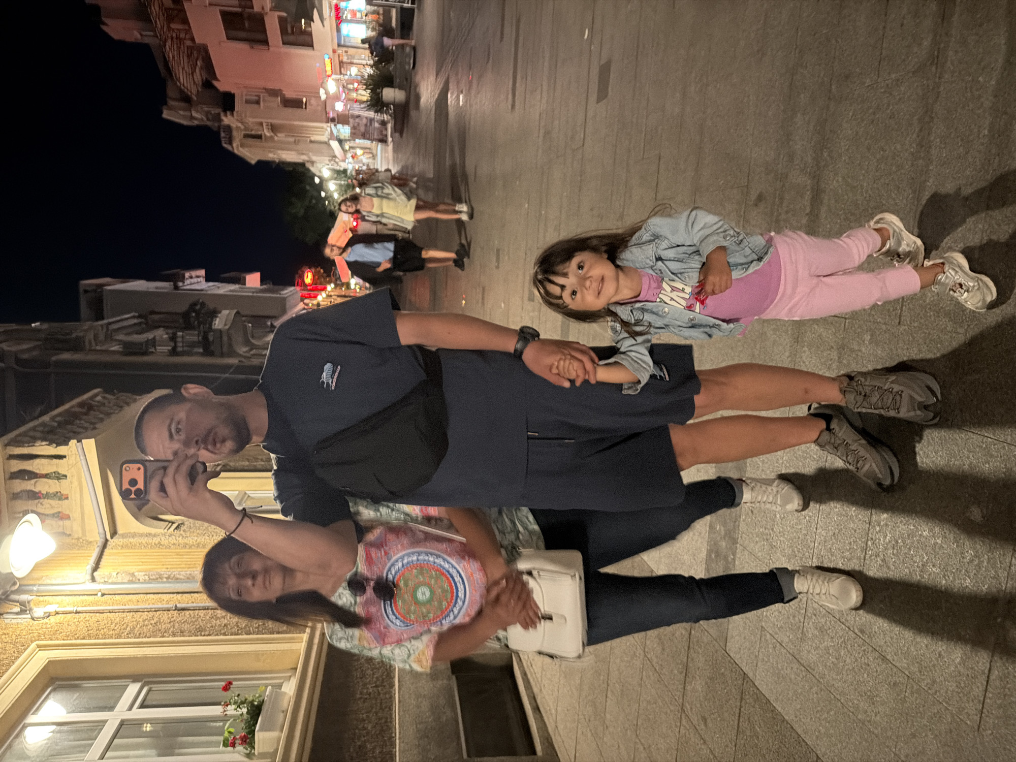 In Burgas, Bulgaria, on a pedestrian street illuminated by streetlights and storefronts, a family is captured in an evening moment. In the foreground, a young girl, about five years old, stands holding the hand of an adult male. She wears a denim jacket over a pink t-shirt and pink pants, with silver sneakers. The man, with short dark hair and a beard, is dressed in a navy blue t-shirt and shorts, with a black fanny pack slung across his chest. He holds a smartphone in his right hand, seemingly taking a photo or video. Behind them, a woman with long brown hair wears a colorful patterned t-shirt and blue jeans, carrying a white crossbody bag. Her arms are crossed, and she looks towards the phone. In the background, other pedestrians stroll along the street, with shops lining both sides. The architecture is a mix of older buildings with balconies and more modern structures. The overall scene depicts a casual outing in a lively urban environment at night.