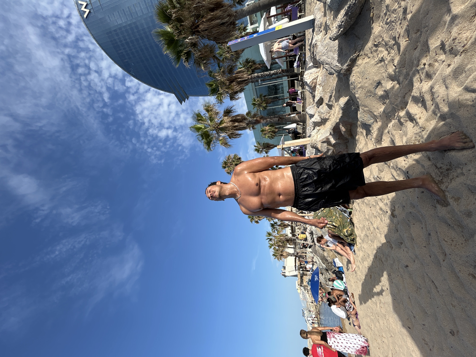 A low-angle, full-shot photograph captures a wet, shirtless man with dark hair standing on a sandy beach in Barcelona, Spain. He is wearing black swim trunks and a silver chain. His face is tilted upwards, eyes closed, and his body is glistening with water, suggesting he has recently emerged from the sea. The man is centrally framed, with his shadow cast long on the sand in the foreground.
Behind him, the beach is populated with other beachgoers lounging on towels, under umbrellas, and near rocky outcrops. Palm trees line the background, with a modern, curved glass building, the W Barcelona Hotel, prominently visible on the right side. The sky is a vibrant blue, dotted with scattered white clouds. The overall impression is a sunny day at a bustling urban beach.